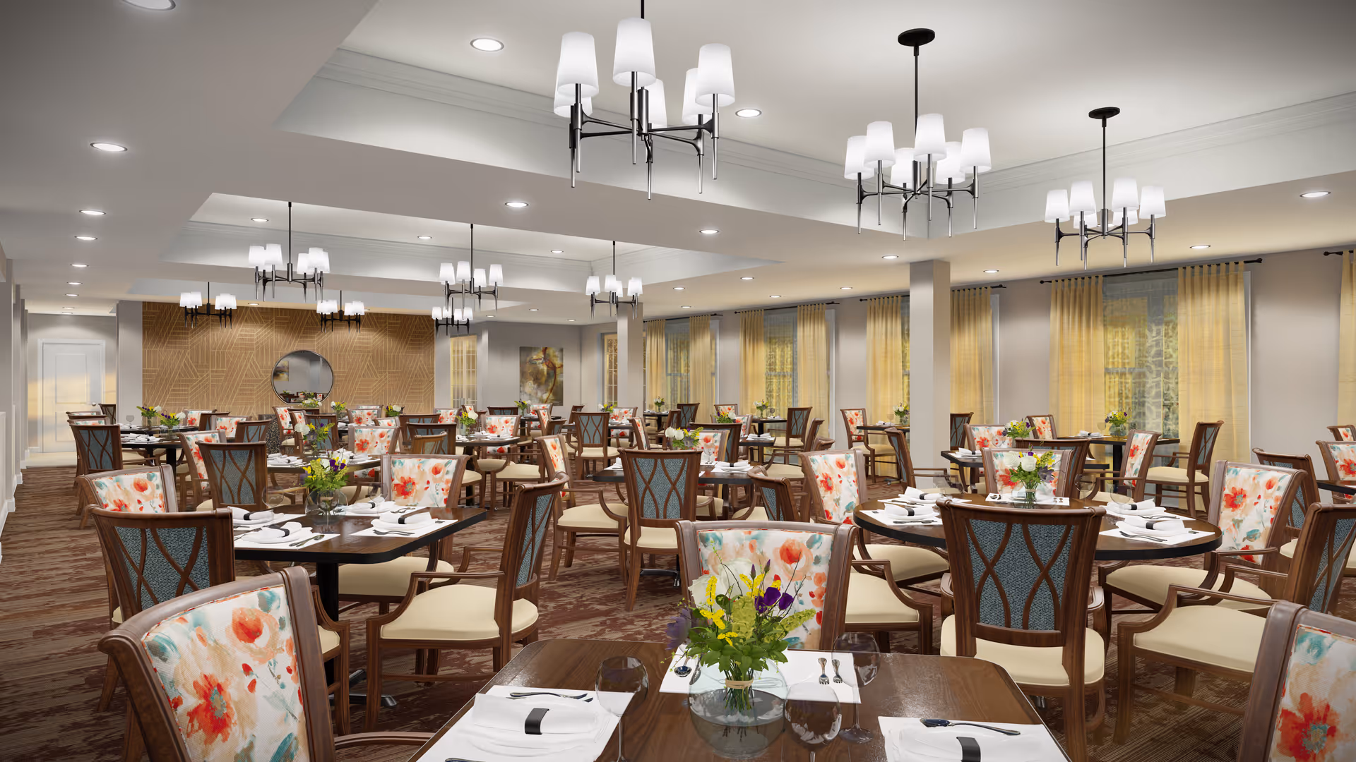 A spacious and elegant dining room with multiple round and square tables set with white napkins, plates, and silverware. The chairs have floral patterned upholstery on the backrests and cream-colored seats. The room features large windows with light yellow curtains, modern chandeliers hanging from the ceiling, and a decorative wall with a round mirror and artwork in the background.