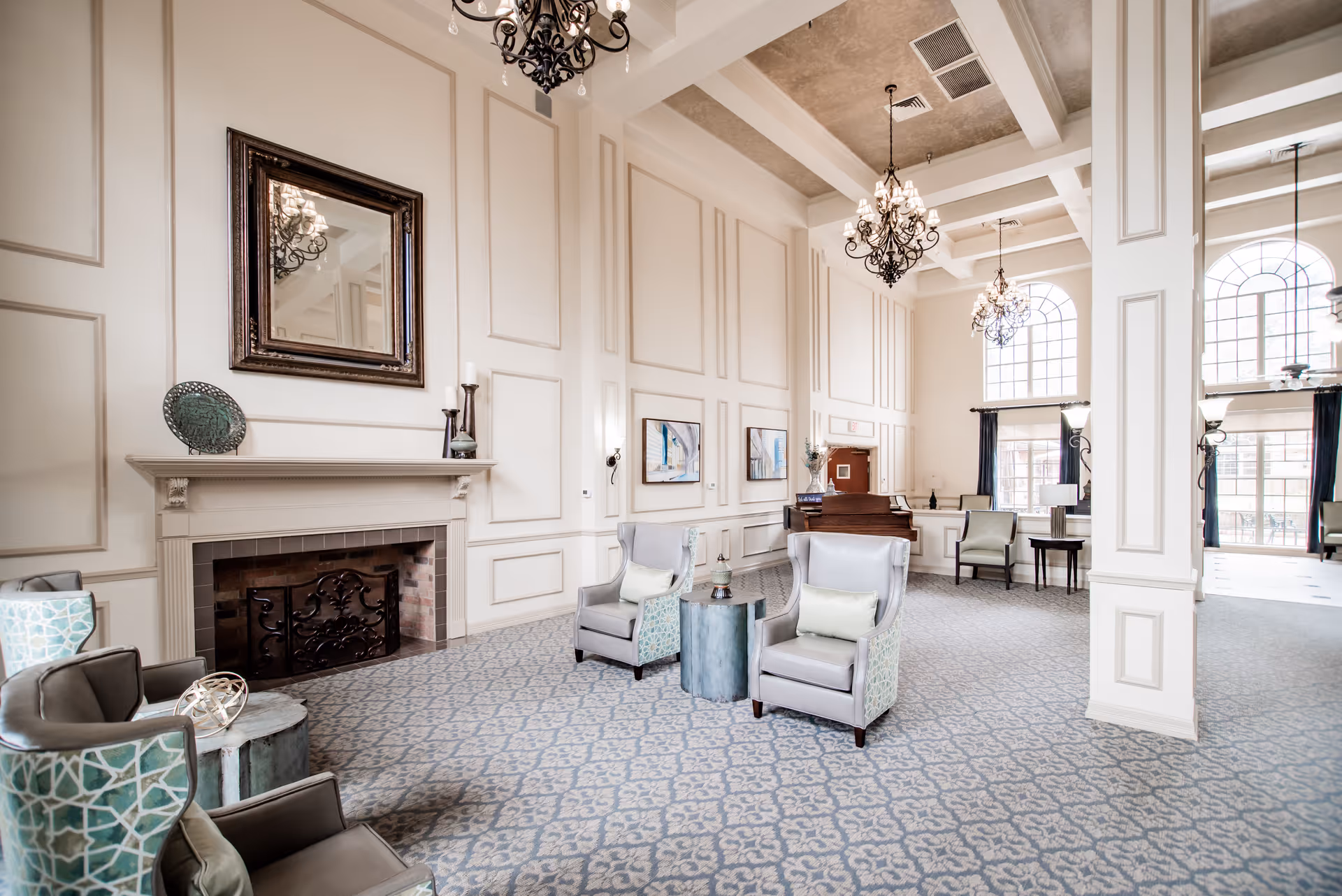 A spacious and elegant living room area with high ceilings, decorative wall paneling, and large windows letting in natural light. The room features a fireplace with a large framed mirror above it, several comfortable armchairs arranged around small tables, chandeliers hanging from the ceiling, and a piano in the background.