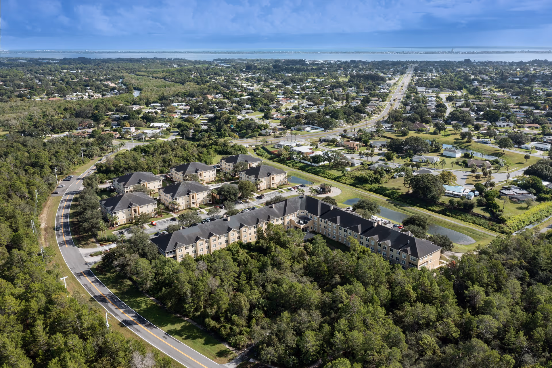 Aerial view of Glenbrooke Senior Living facility surrounded by trees and greenery, with multiple buildings and parking areas visible. The facility is located near a curved road and a larger residential neighborhood extends into the distance under a partly cloudy sky.