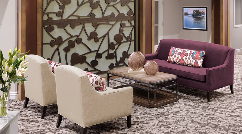 A cozy seating area in a senior living facility featuring a purple sofa with a colorful patterned pillow, two beige armchairs with matching pillows, a wooden coffee table with decorative vases, a floral-patterned carpet, a decorative wooden wall panel, and a framed landscape painting on the wall.
