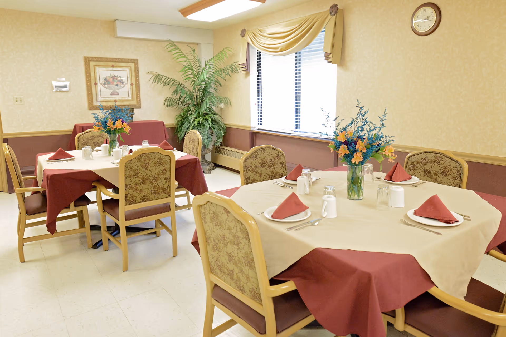 A dining room with two tables covered in beige and burgundy tablecloths, each set with plates, utensils, glasses, and red folded napkins. There are floral centerpieces on each table, upholstered chairs around the tables, a large window with blinds and a valance, a framed picture on the wall, a clock, and a large potted plant in the corner.