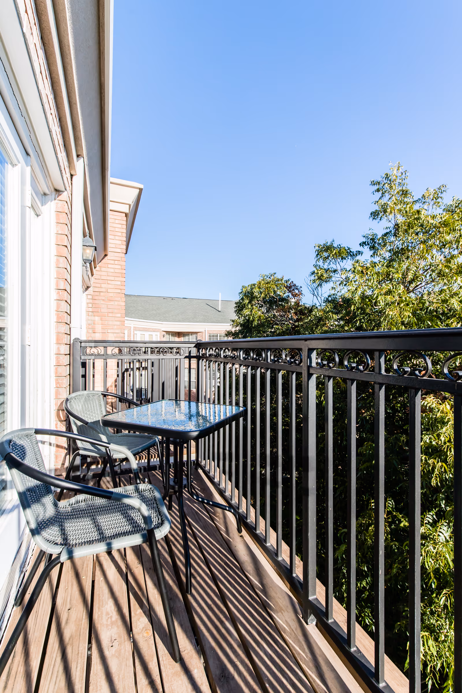 Narrow balcony with a small glass-top table and two metal chairs, metal railing, trees and a neighboring building under a clear blue sky.