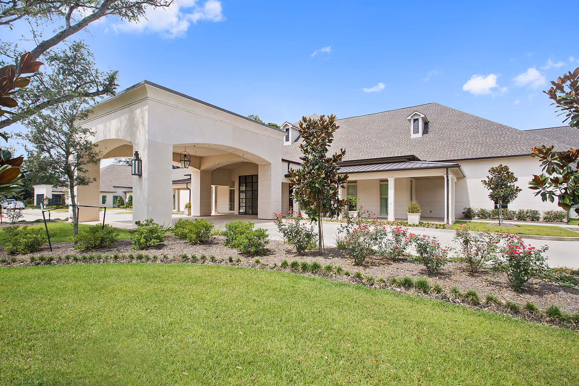 Exterior view of a senior living facility with a covered entrance, manicured lawn, flowering bushes, and trees under a blue sky with scattered clouds.