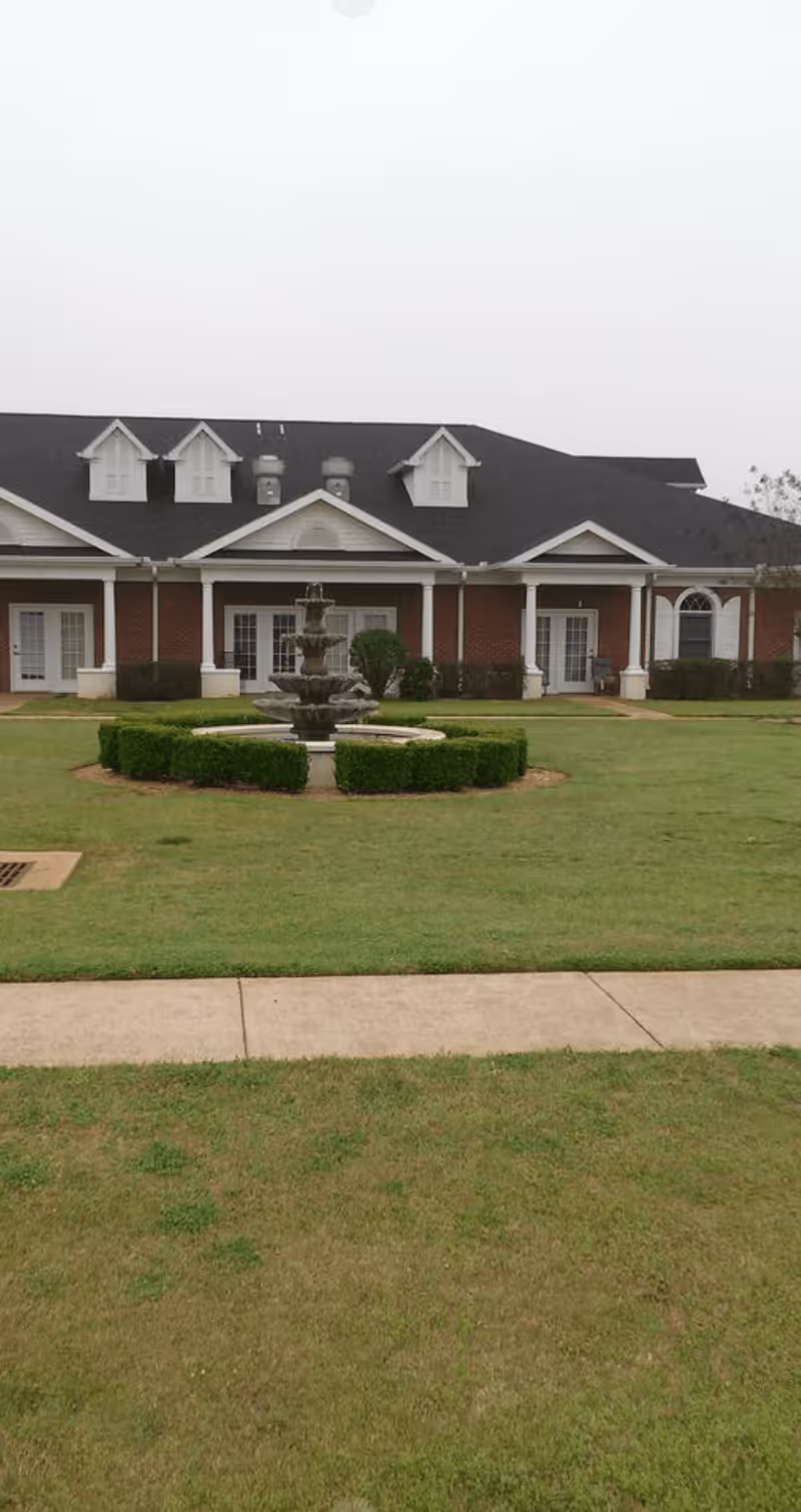 A large brick building with white trim and multiple dormer windows on the roof, surrounded by a well-maintained lawn with a circular hedge and a tiered water fountain in the center. A concrete walkway runs in front of the lawn.