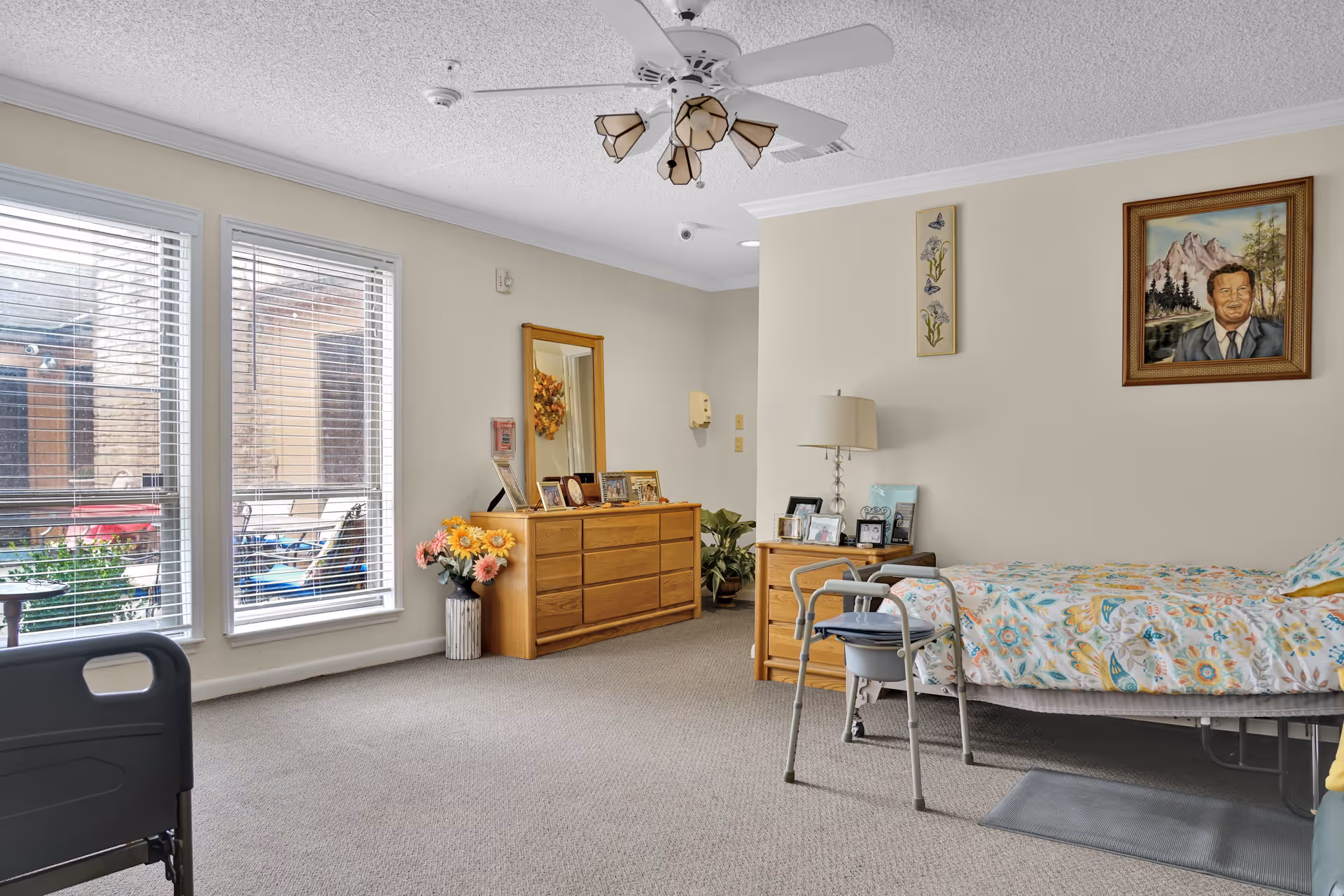 A well-lit bedroom in a senior living facility featuring a single bed with a colorful floral bedspread, a bedside table with a lamp and framed photos, a wooden dresser with a mirror and more framed photos, a walker positioned near the bed, large windows with blinds letting in natural light, and a ceiling fan with lights. There is a painting of a man on the wall and a vase with flowers near the dresser.