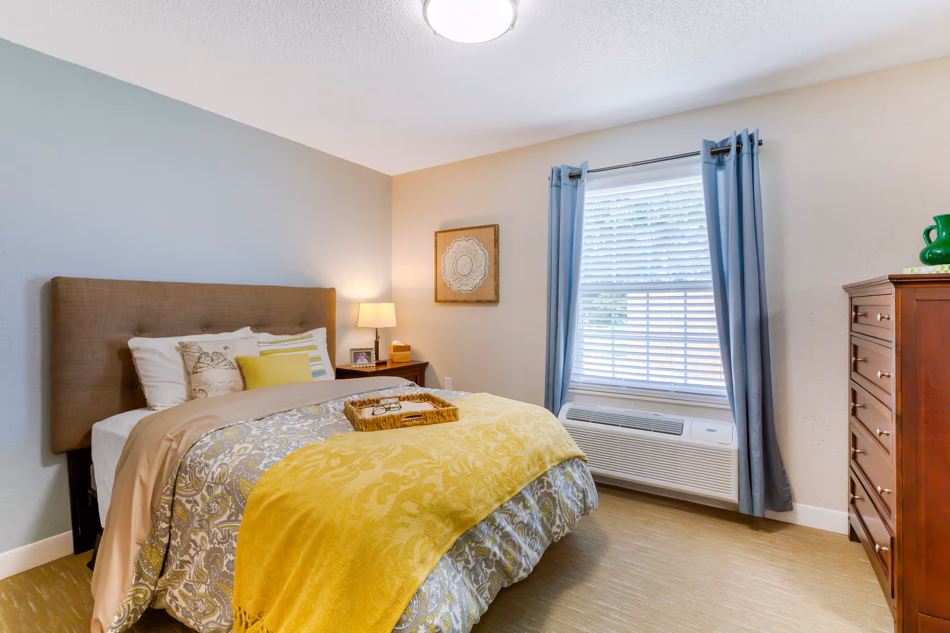 A cozy bedroom with a bed featuring a brown upholstered headboard, patterned bedding with a yellow throw blanket, and several pillows. A wooden nightstand with a lamp, picture frame, and tissue box is beside the bed. There is a window with blue curtains and white blinds, and a wooden dresser with a green pitcher on top. The walls are painted in light neutral tones.