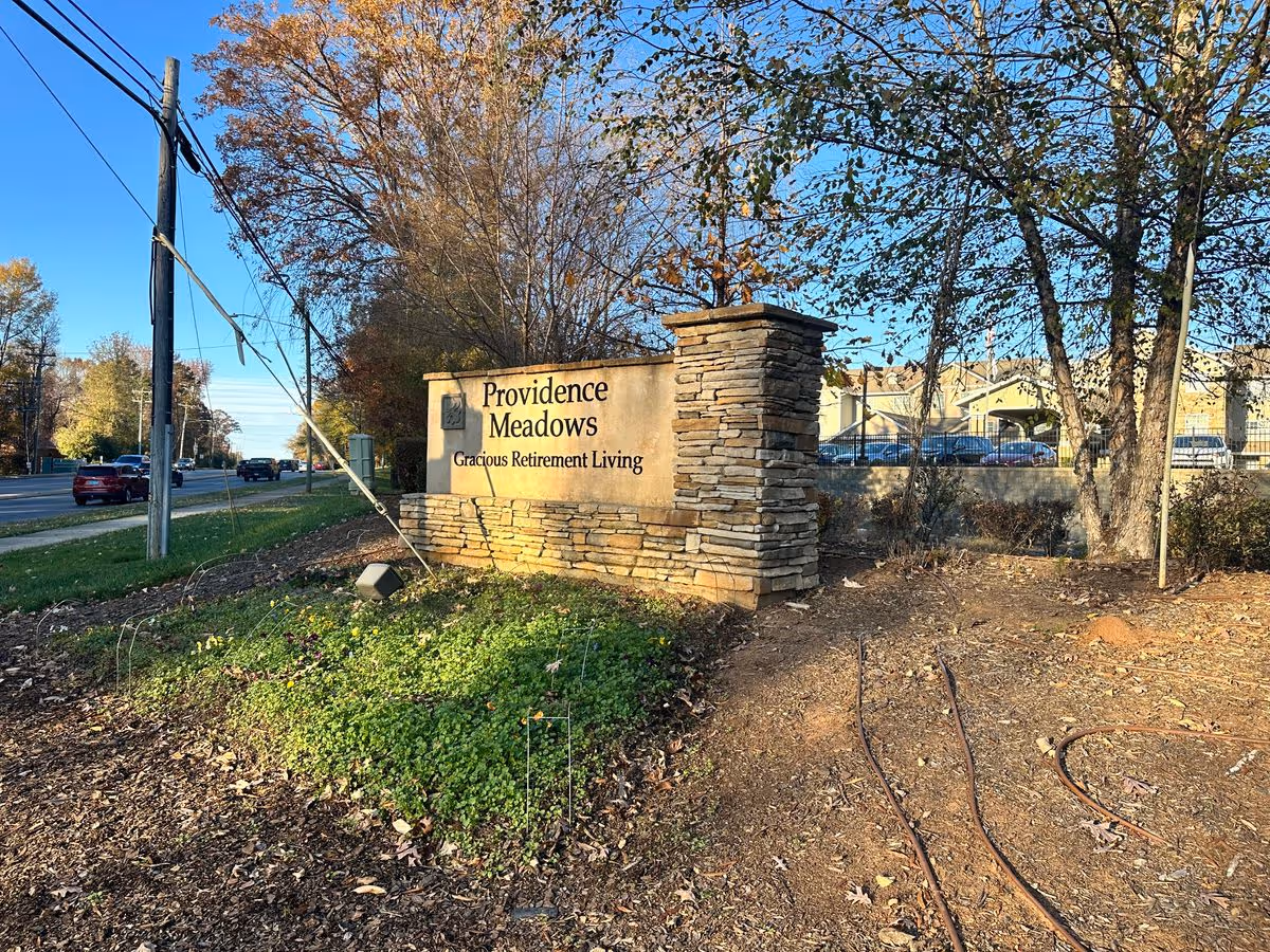 Stone entrance sign reading "Providence Meadows Gracious Retirement Living" at the roadside entrance with trees and the facility buildings visible behind it.