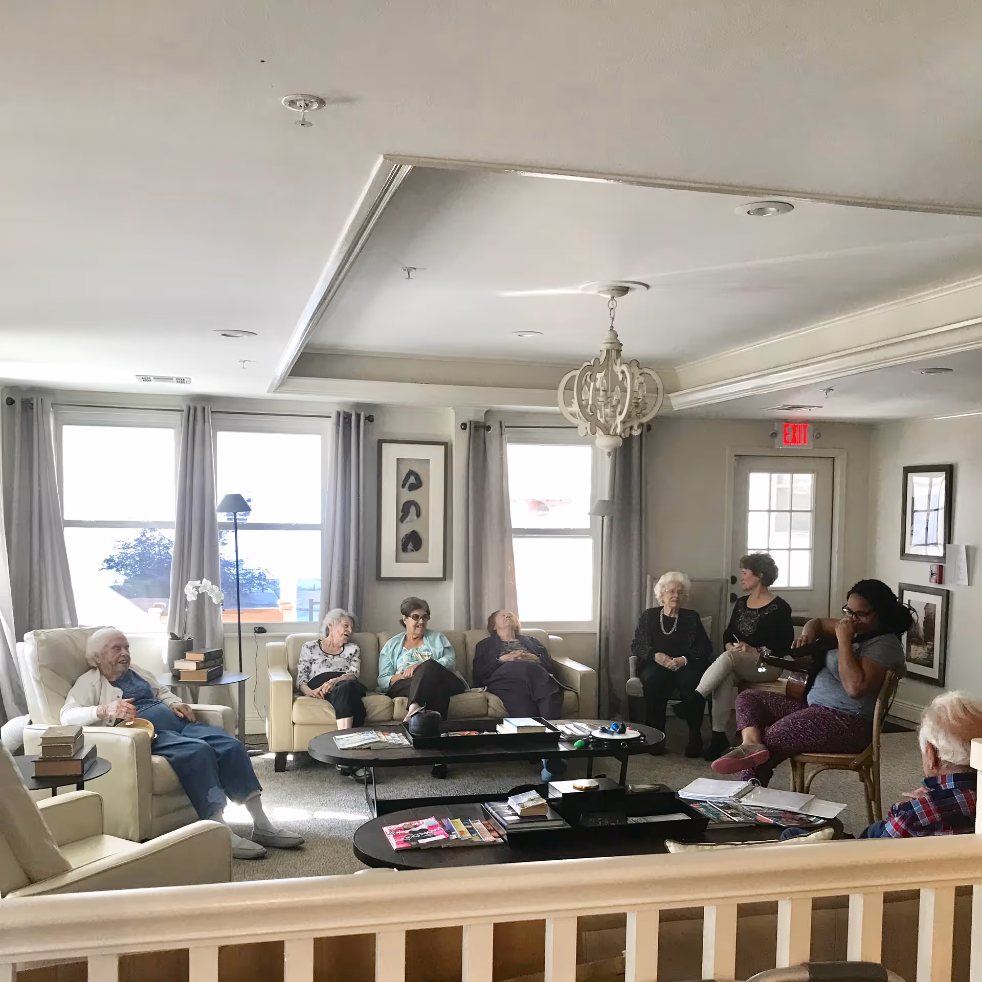 Bright communal living room with seniors seated on sofas and chairs around coffee tables under a chandelier.