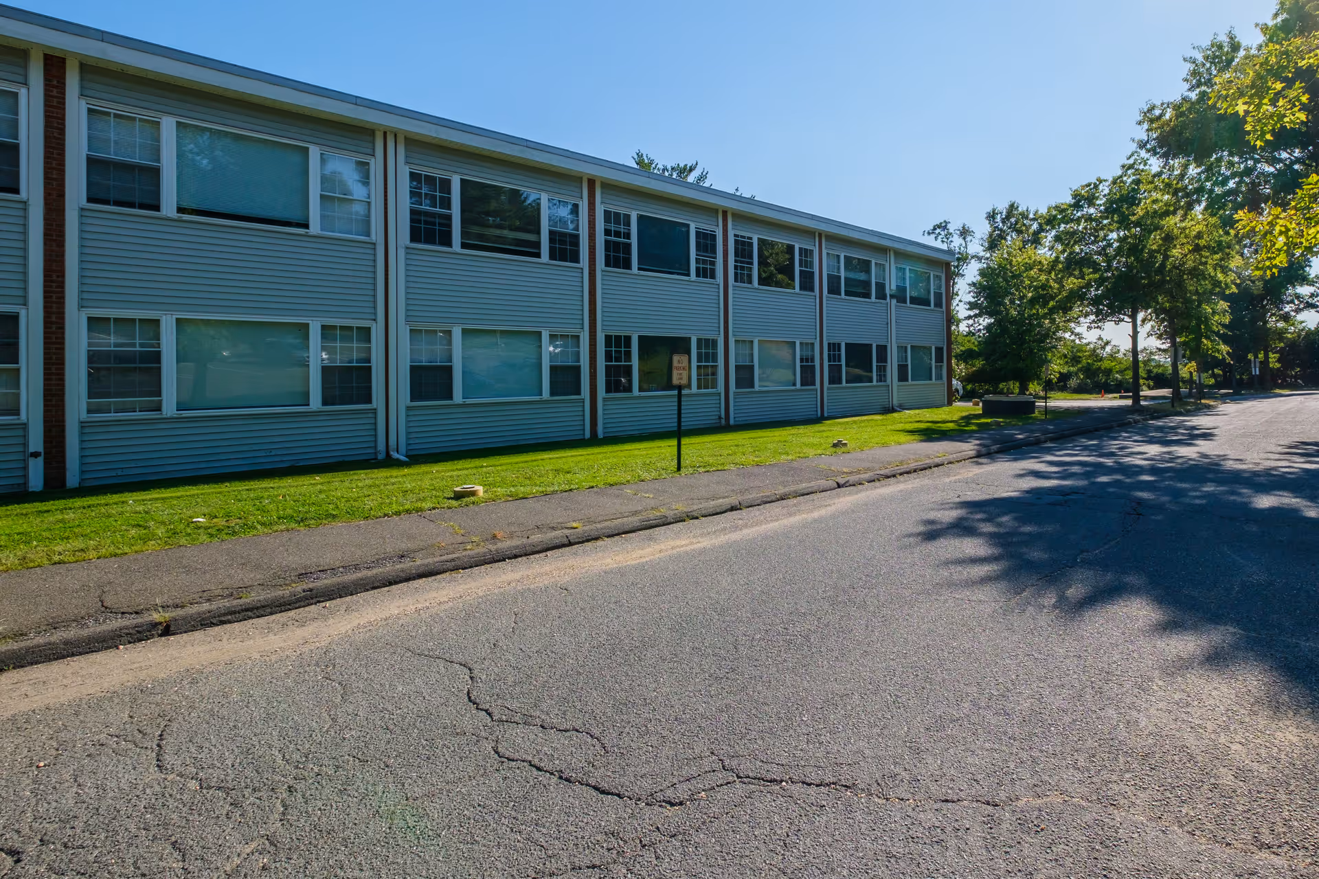 Exterior view of a two-story building with multiple windows, a sidewalk, and a paved road in front. There is green grass and trees along the side of the building under a clear blue sky.