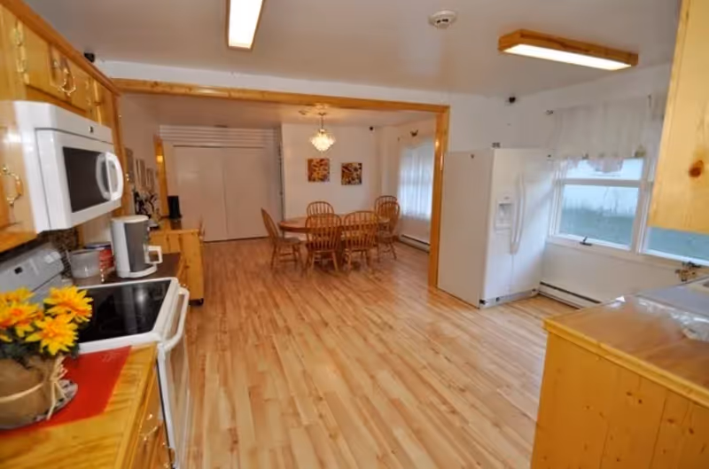 A spacious kitchen and dining area with wooden cabinets, a white microwave, stove, refrigerator, and a dining table with six wooden chairs. The floor is light wood, and there are two windows with sheer white curtains allowing natural light into the room.
