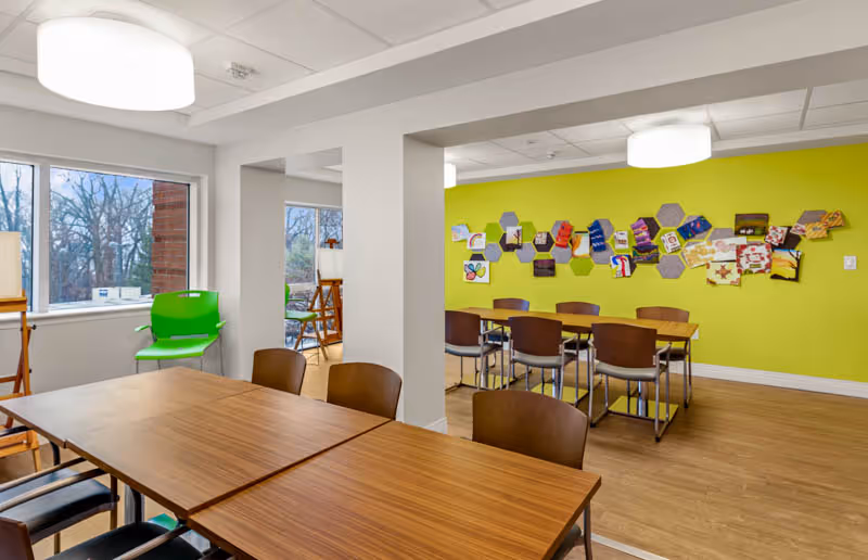 A bright activity room with wooden tables and chairs arranged for group seating. The walls are painted white and lime green, with colorful artwork displayed on the green wall. Large windows let in natural light and show trees outside. The room has a clean, modern look with ceiling lights and wooden flooring.
