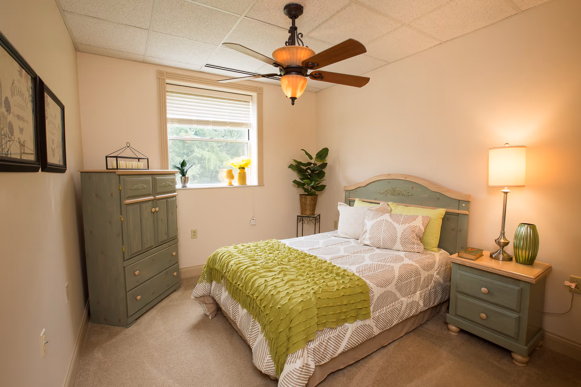 A cozy bedroom with a bed featuring a green and white patterned bedspread and green throw blanket. The room has a green wooden headboard, matching nightstand with a lamp and decorative vase, and a green wooden dresser near a window with blinds. There are framed pictures on the wall and a potted plant in the corner.
