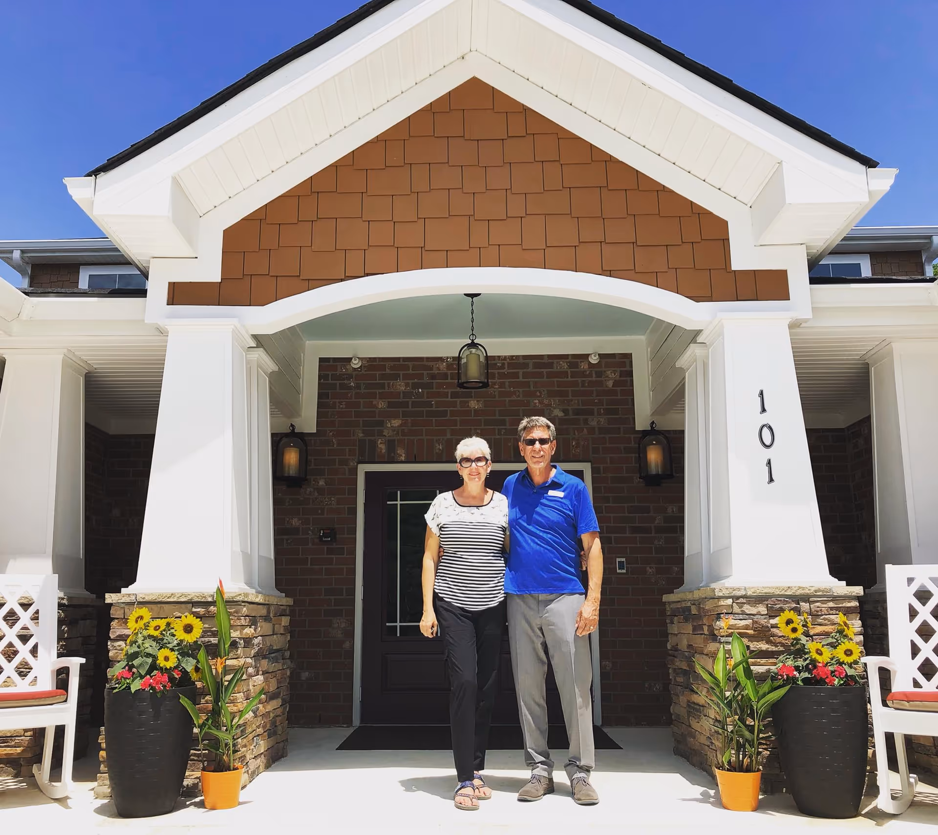 An elderly woman and man standing arm in arm in front of the entrance to a building with brick walls, white columns, and a peaked roof. There are large potted plants with sunflowers on either side of the entrance, and two white chairs with red cushions are partially visible. The building number 101 is displayed on one of the columns.