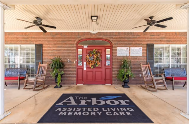 Entrance of The Arbor Assisted Living and Memory Care facility with a red door decorated with a wreath, two rocking chairs and benches on either side, potted plants, ceiling fans, and a large black mat with the facility's name printed on it.