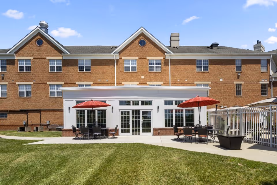 Brick senior living building with a patio area featuring tables, chairs, and red umbrellas on a grassy lawn.
