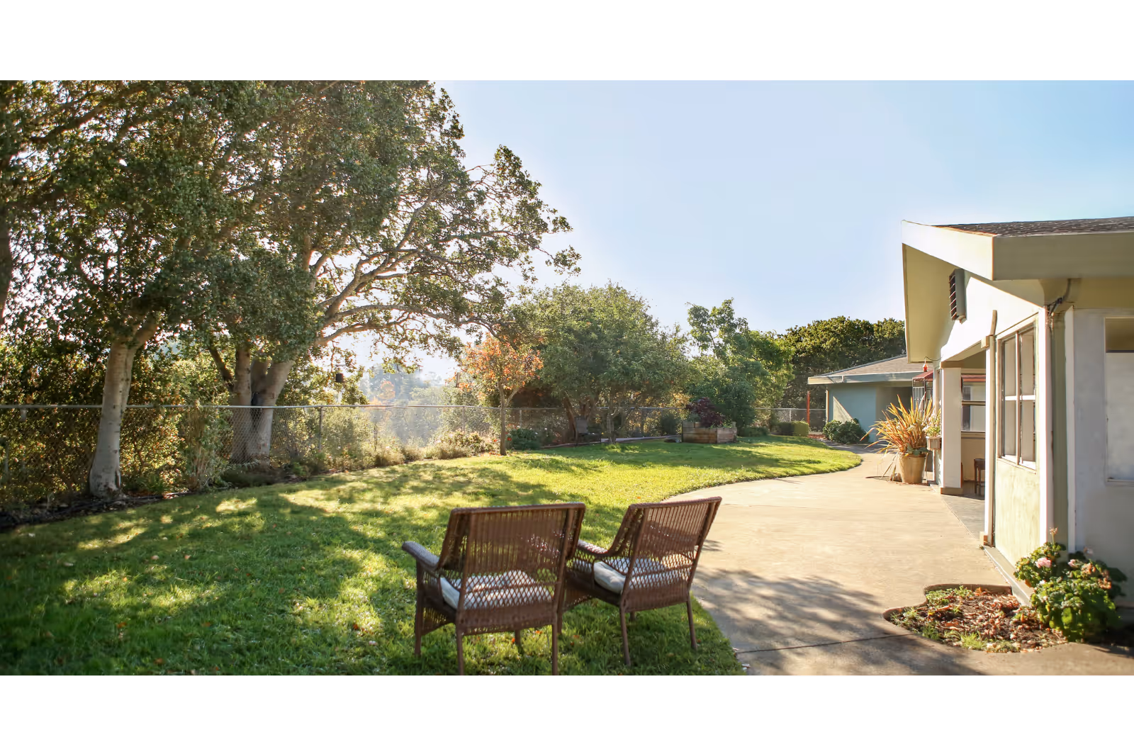 Sunlit outdoor patio and lawn with two wicker chairs facing a grassy yard and trees beside a single-story building.