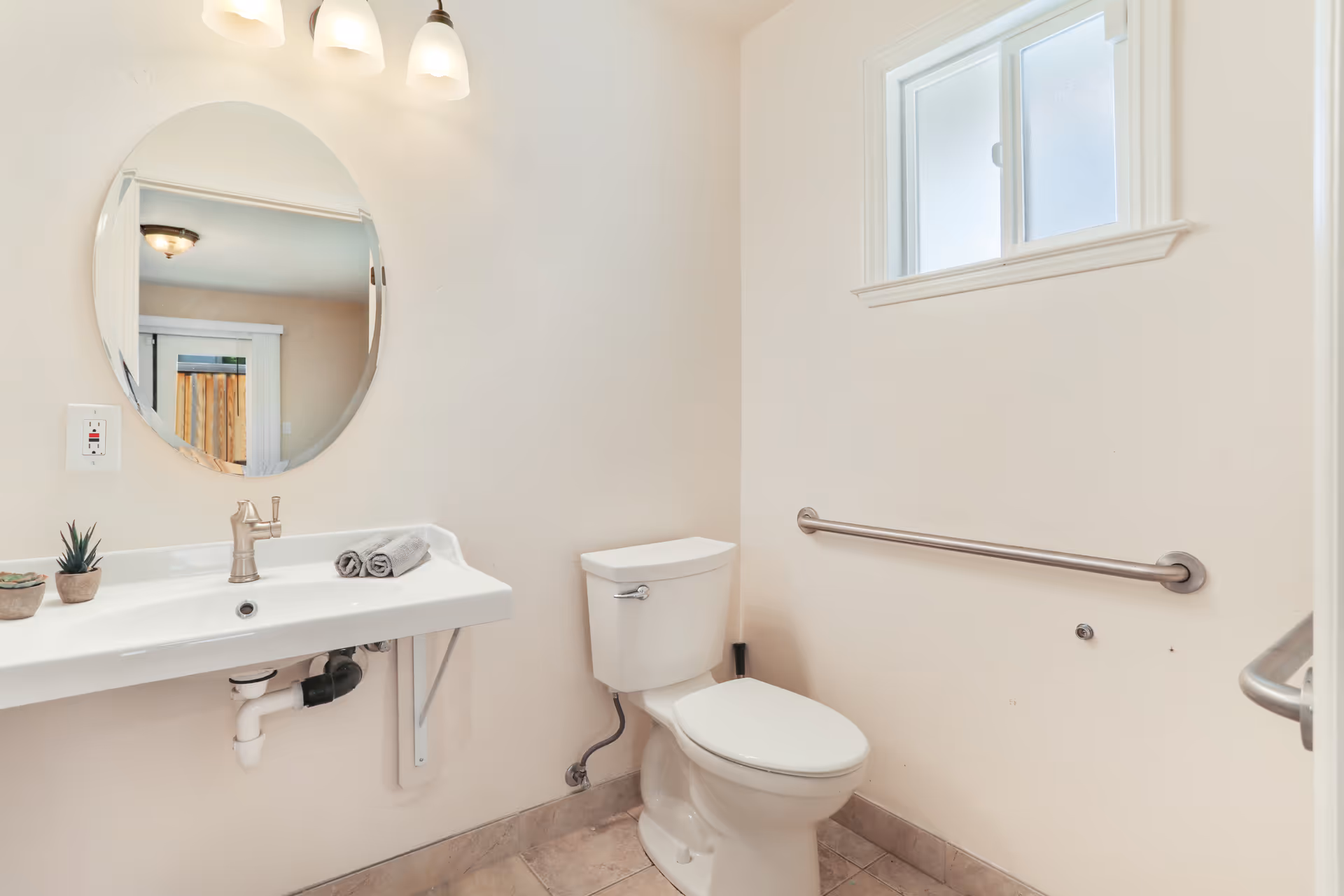 A clean and simple bathroom with a white toilet, a wall-mounted white sink with a silver faucet, an oval mirror above the sink, two small potted plants on the sink, two rolled gray towels, a small frosted window, and a metal grab bar on the wall next to the toilet.
