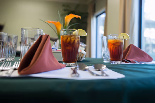 Close-up view of a dining table set with glasses of iced tea garnished with lemon wedges, folded brown cloth napkins, silverware, and a green tablecloth. In the background, there is a flower centerpiece and windows letting in natural light.