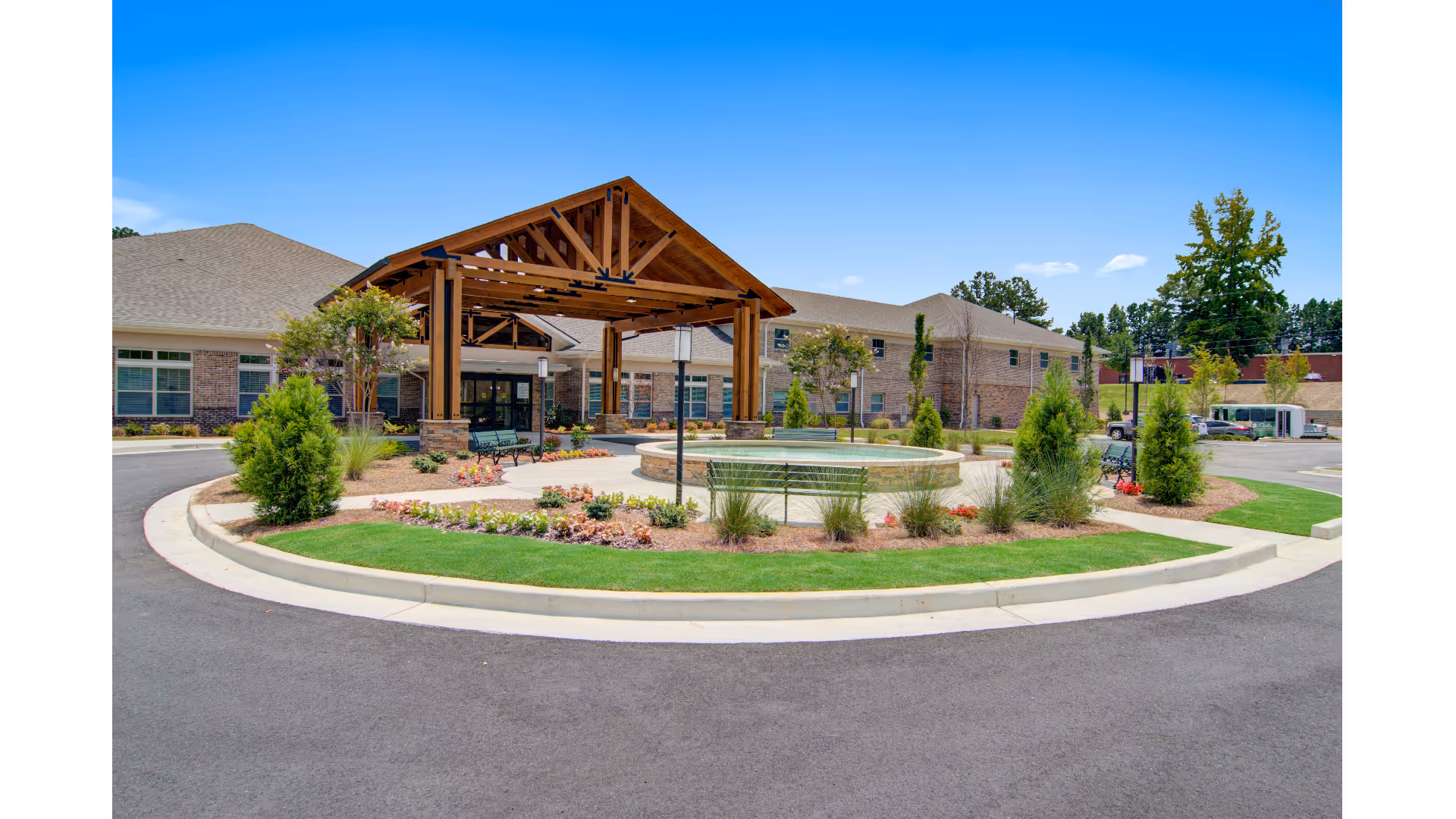 Exterior view of Prema at Suwanee Creek senior living facility showing a circular driveway with landscaped greenery, benches, a round water feature, and a large wooden entrance canopy under a clear blue sky.