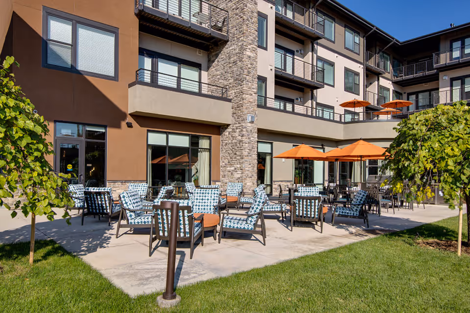 Outdoor patio with patterned lounge chairs, orange umbrellas, and tables in front of a multi-story senior living building.