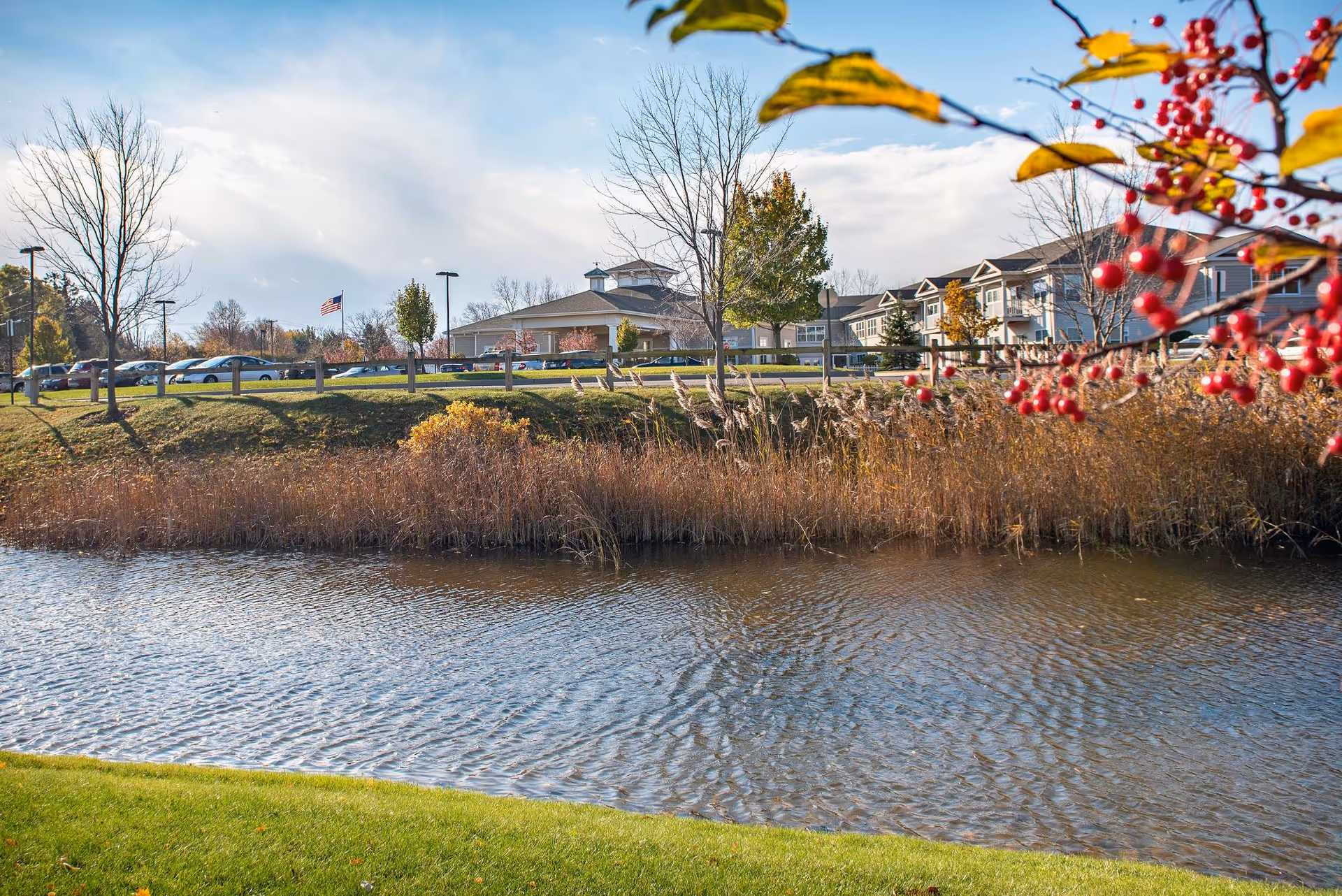 View of a senior living facility named The Addison of Erie Station across a small body of water with reeds and grass in the foreground. The building is surrounded by trees, some with autumn foliage, and a parking lot with cars. An American flag is visible near the building under a partly cloudy sky.