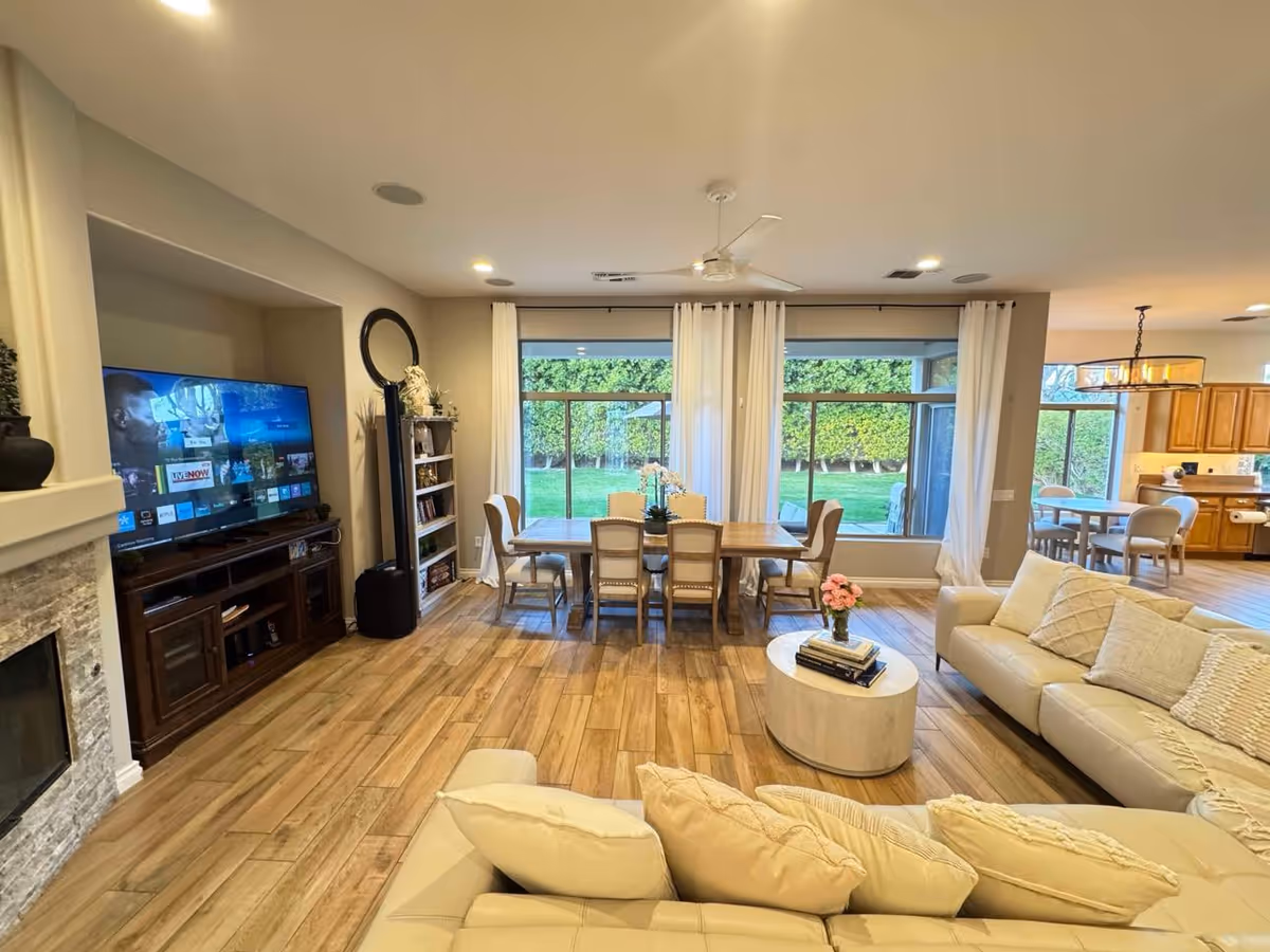 A spacious and well-lit living and dining area with wooden flooring. The living room features a large beige sectional sofa with multiple cushions, a round white coffee table with books and a flower vase, and a flat-screen TV on a dark wooden stand next to a stone fireplace. The dining area has a wooden table with six chairs and large windows with white curtains overlooking a green garden. In the background, a kitchen with wooden cabinets and a dining nook is visible.