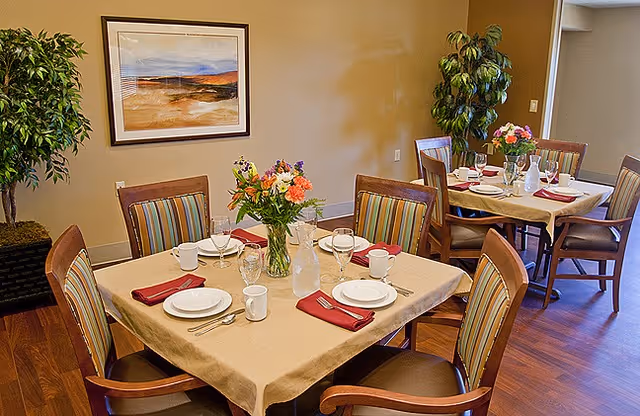 A dining area with two square tables covered with beige tablecloths, each set for four people with white plates, cups, silverware, and red napkins. Each table has a vase with colorful flowers. The room has wooden floors, striped cushioned chairs, a large potted plant, and a framed landscape painting on the wall.