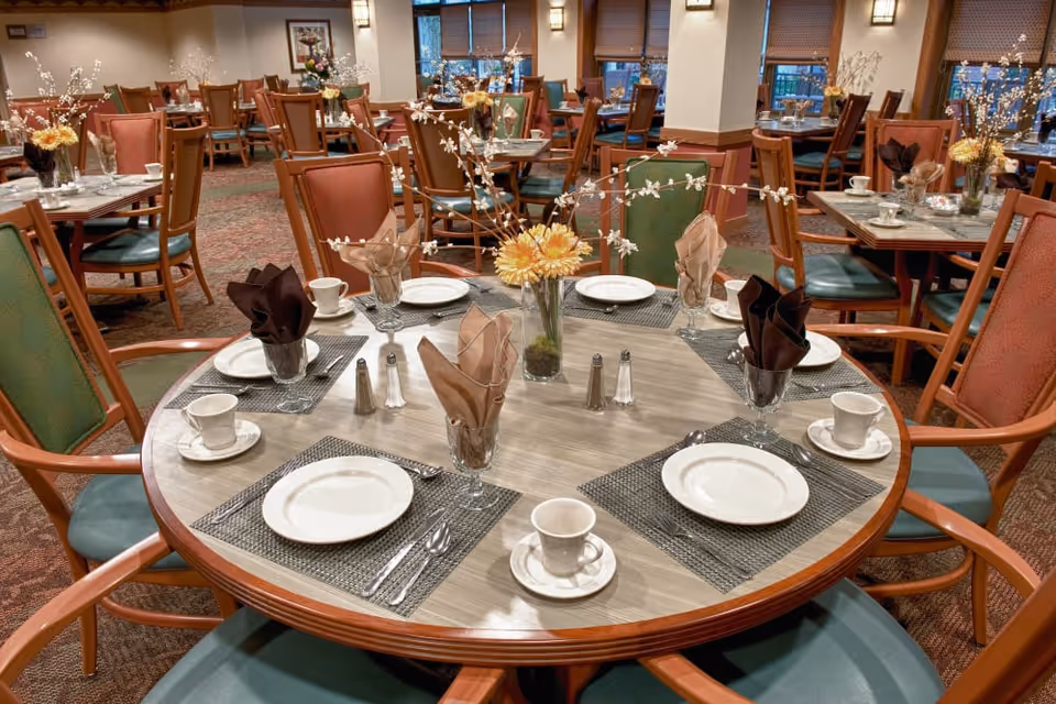 A dining room with round tables set with plates, cups, folded napkins, and floral centerpieces.