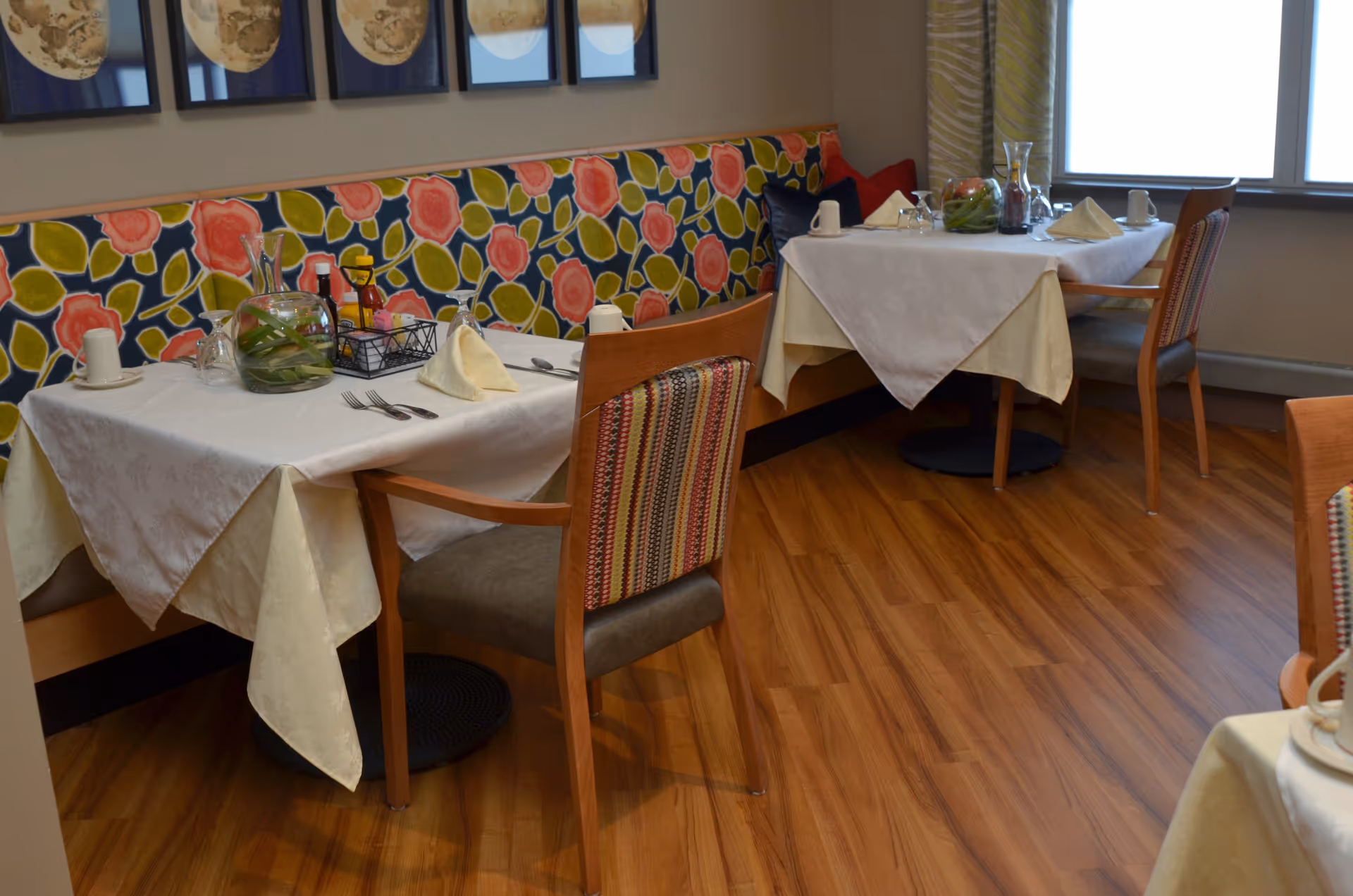 A dining area with two tables covered with white tablecloths and set with cups, napkins, and utensils. The tables are accompanied by wooden chairs with patterned upholstery. The back wall features a colorful floral-patterned cushioned bench, and there are framed artworks hanging above it. The floor is wooden, and a window is visible in the background.