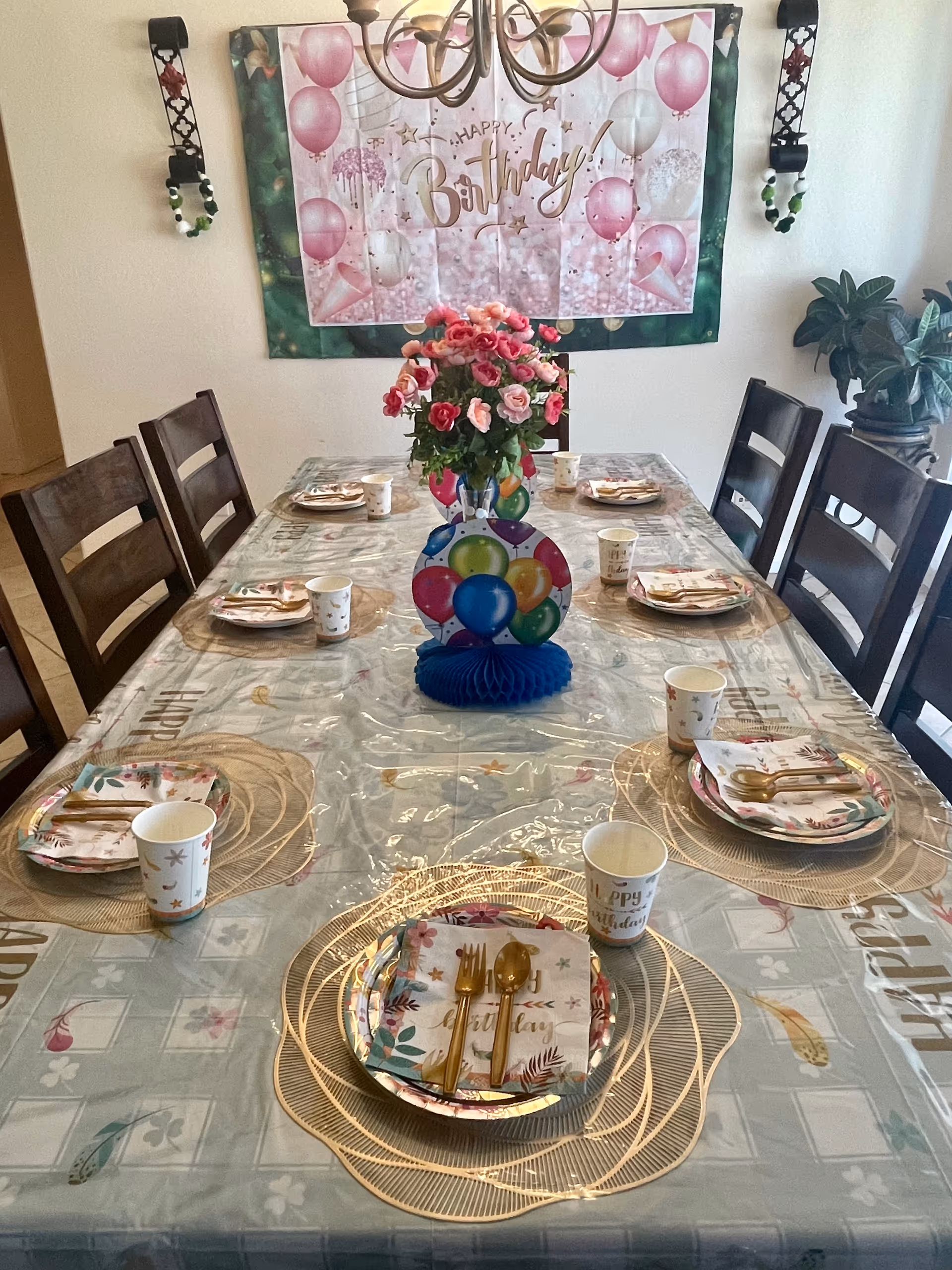 A dining table set for a birthday celebration with six place settings. Each setting has a floral plate, a napkin with a birthday design, gold-colored cutlery, and a paper cup. The table is covered with a decorative tablecloth and a clear plastic cover. In the center of the table is a bouquet of pink and red flowers and a colorful balloon decoration. On the wall behind the table is a large 'Happy Birthday' banner with pink and white balloons. The room has wooden chairs around the table and a plant in the corner.