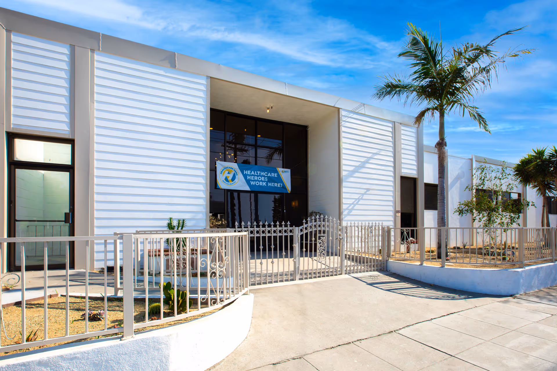 Exterior view of a white building with a gated entrance and palm trees under a blue sky. A sign near the entrance reads 'Healthcare Heroes Work Here!'