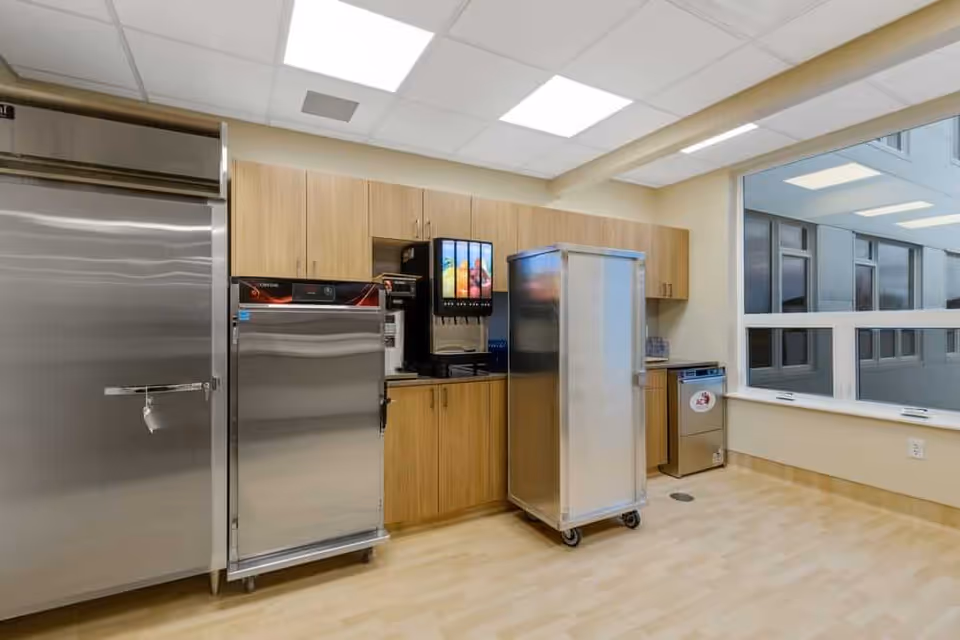 A clean and modern kitchen area with stainless steel appliances including a large refrigerator, a smaller refrigerator, a beverage dispenser, and a mobile storage cart. Light wood cabinets line the back wall, and there is a large window on the right side letting in natural light.