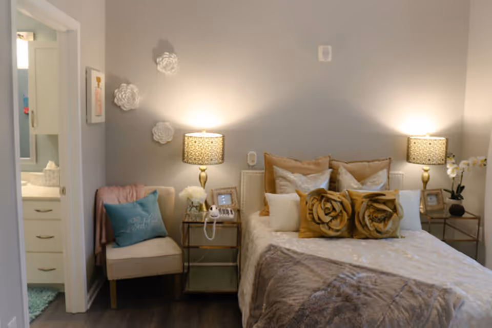 A cozy bedroom with a neatly made bed featuring decorative pillows, including two large rose-shaped pillows. There are two bedside tables with matching lamps, a small chair with a teal pillow and a pink throw blanket, and wall decorations shaped like white flowers. An open door reveals a glimpse of a bathroom with white cabinetry.