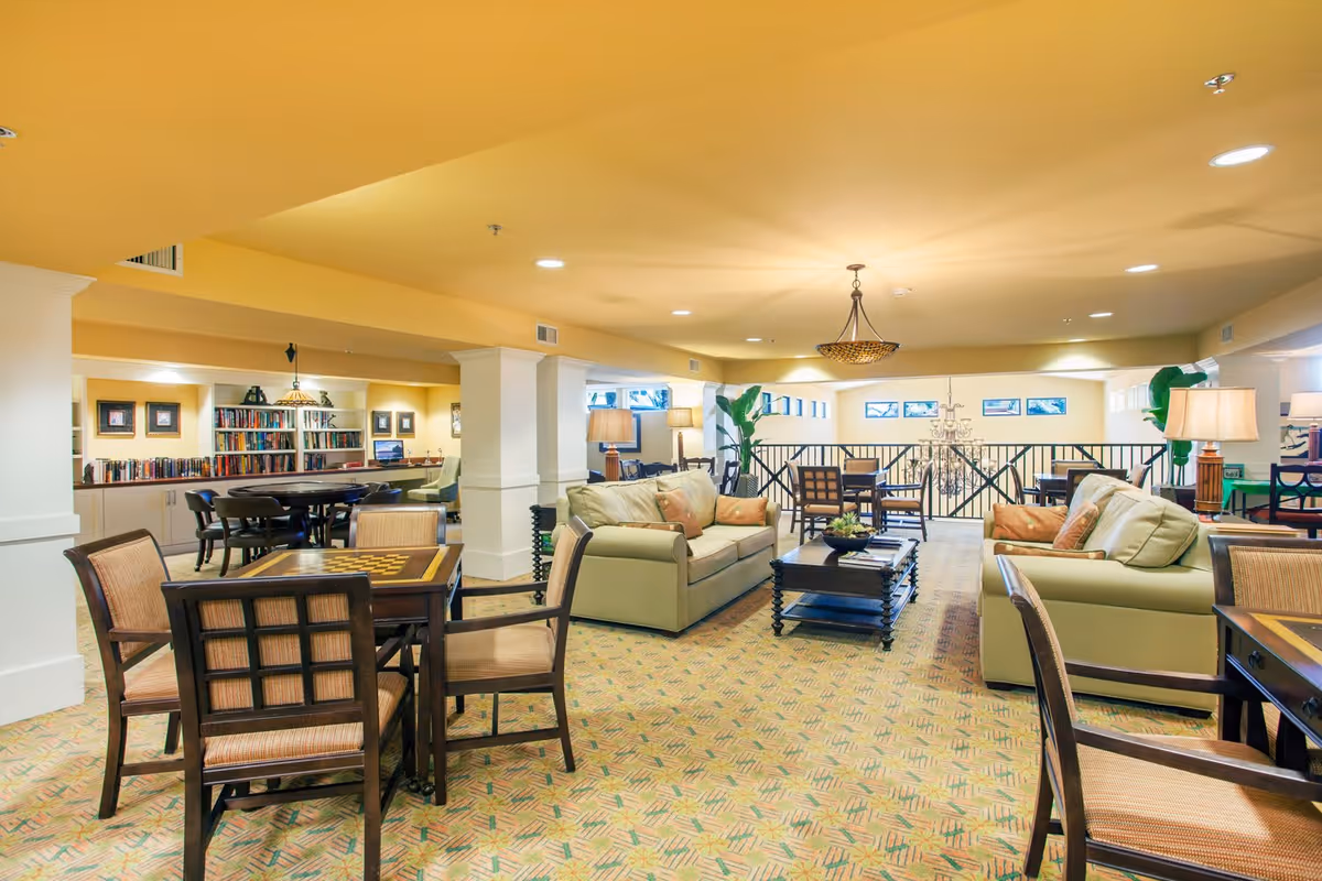 A spacious and well-lit common area in a senior living facility featuring multiple seating arrangements including sofas with cushions, wooden chairs, and tables. The room has a warm yellow ceiling, carpeted floor with a patterned design, bookshelves filled with books, and several lamps providing ambient lighting. There are decorative plants and framed pictures on the walls, creating a cozy and inviting atmosphere.