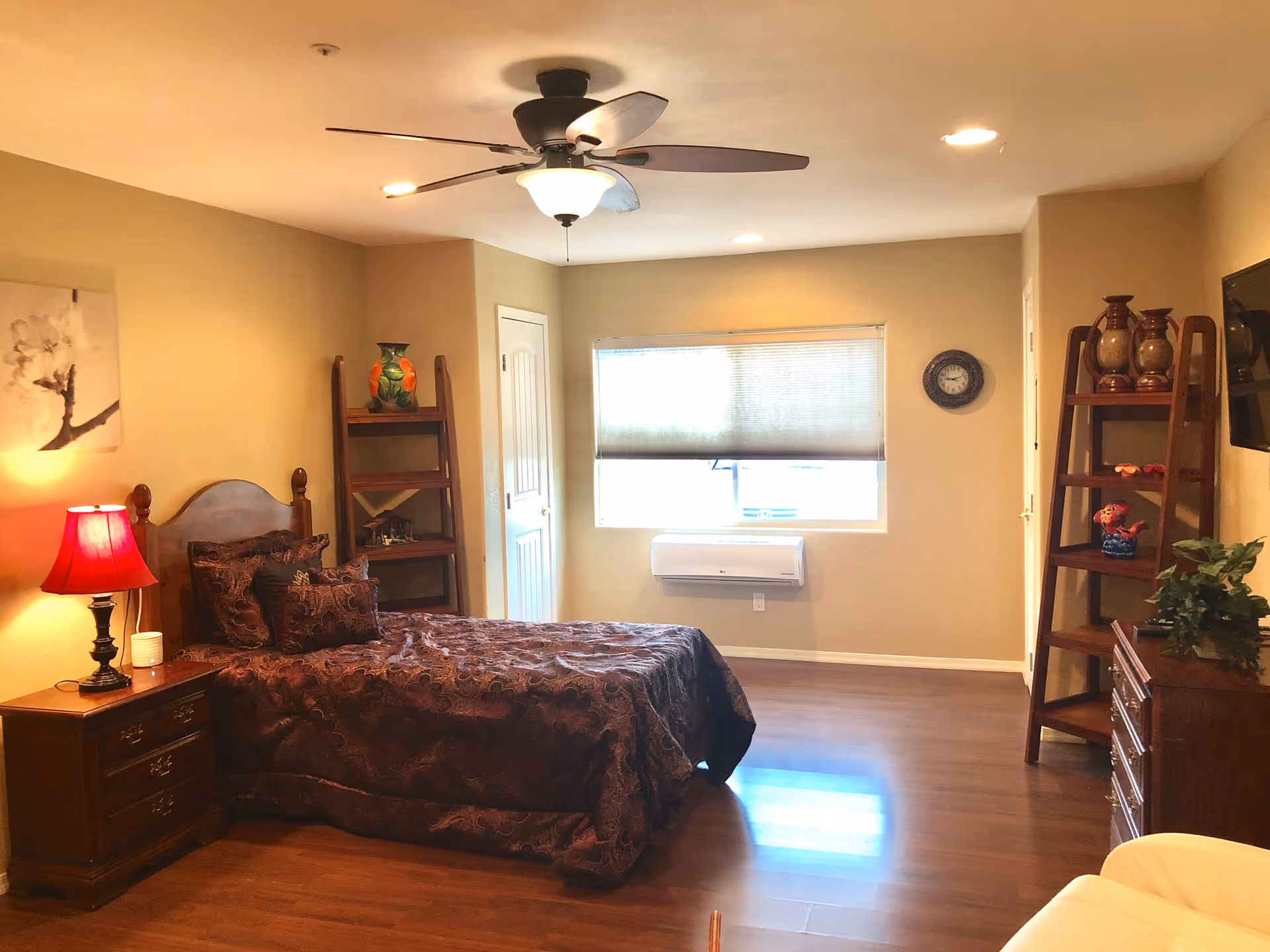 A cozy bedroom in an assisted living facility featuring a wooden bed with a dark patterned bedspread and matching pillows. There is a wooden nightstand with a red lamp and a small white item on it. The room has wooden flooring, beige walls, a ceiling fan with lights, and a window with a partially closed blind. Two wooden ladder-style shelves hold decorative items, and a wall clock is mounted near the window. A flat-screen TV is mounted on the wall opposite the bed, and a wooden dresser with plants and decorative items is visible.