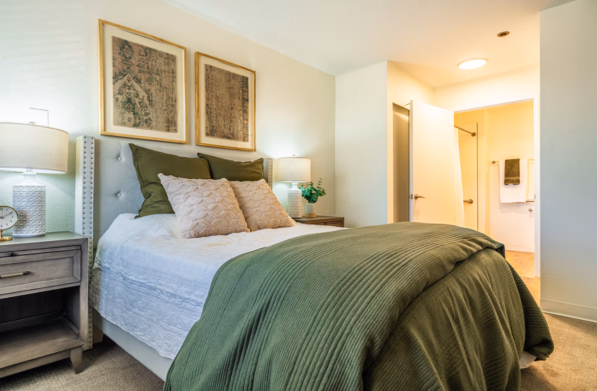 A cozy bedroom with a neatly made bed featuring white and green bedding, two framed artworks above the headboard, two bedside tables each with a lamp, and an open door leading to a bathroom with towels hanging on the wall.