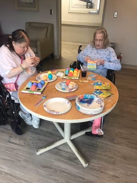 Two elderly women sitting in wheelchairs at a round wooden table engaged in decorating colorful Easter eggs. The table has several plates with painted eggs, egg dye kits, and other decorating supplies. The room has a wooden floor and neutral-colored walls with a chair and hallway visible in the background.