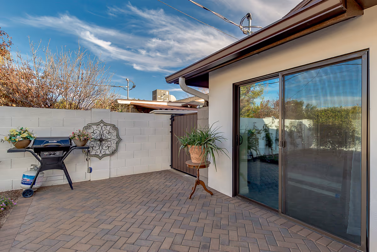 Outdoor patio area with brick paver flooring, a sliding glass door leading inside, a small table with a potted plant, a wall-mounted decorative metal piece, and a gas grill with flower pots on either side. The area is enclosed by a white block wall and a wooden gate.