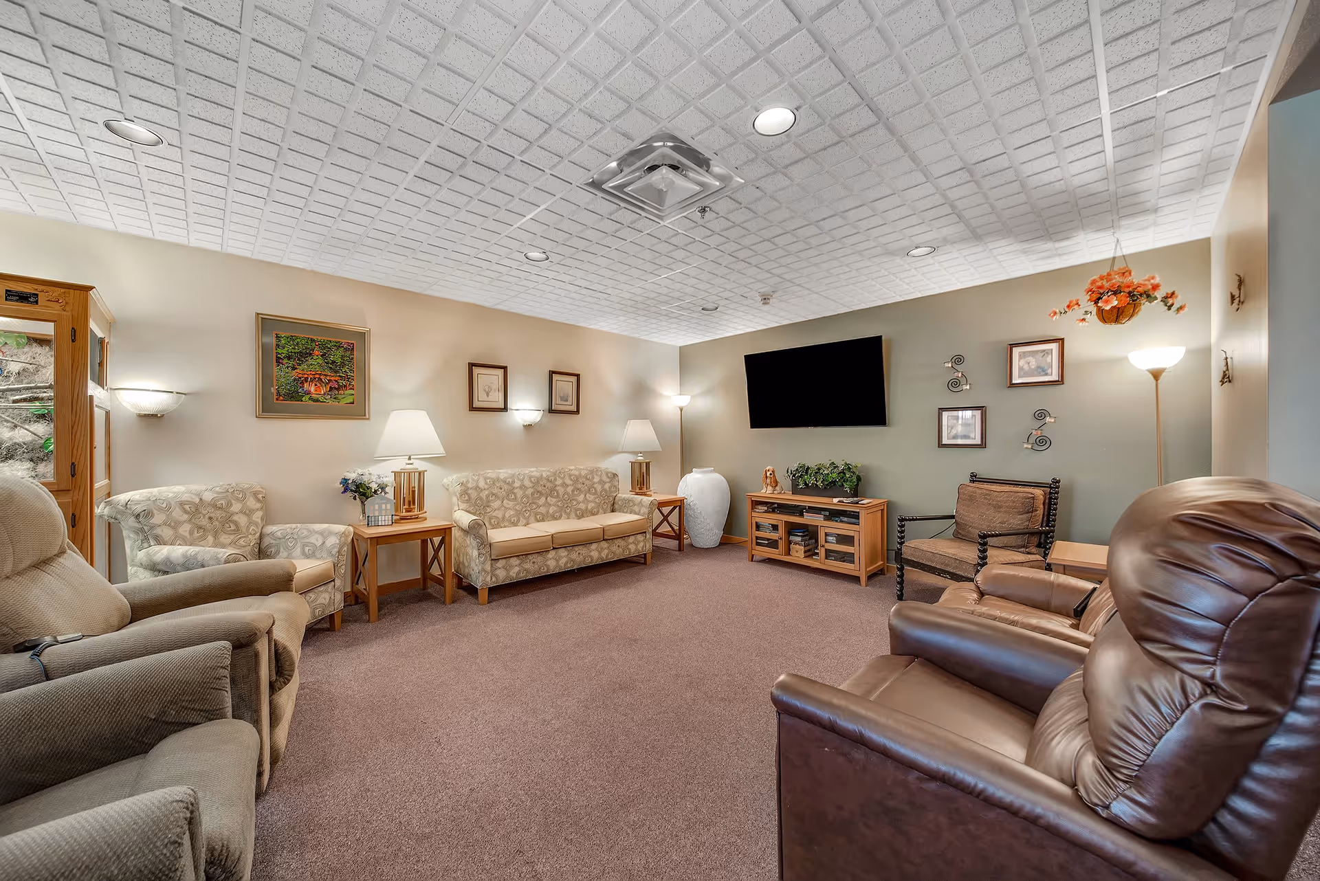 A cozy living room with a variety of seating options including two beige patterned armchairs, a matching loveseat, a brown leather recliner, and a wooden framed chair with a cushion. The room has a carpeted floor, a textured white ceiling with recessed lighting, and walls decorated with framed pictures and a hanging flower basket. A flat-screen TV is mounted on a green accent wall above a wooden media console with decorative items and books.
