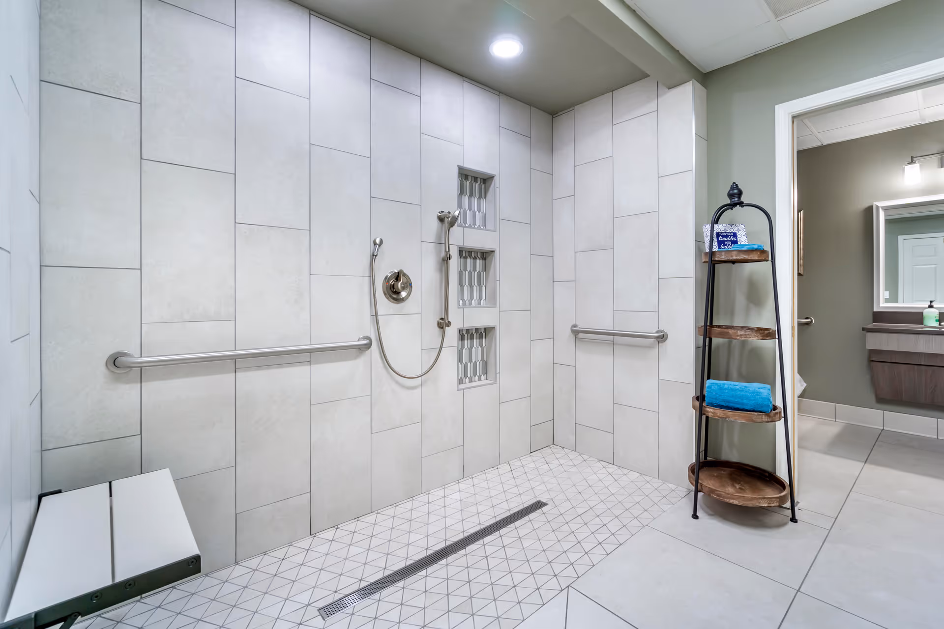 A modern, accessible shower area with large light gray tiles on the walls and smaller patterned tiles on the floor. The shower features a handheld showerhead, built-in wall niches with decorative tiles, and horizontal grab bars. There is a fold-down bench on the left side and a black metal and wood shelving unit with towels and toiletries on the right. The shower opens into a bathroom area with a sink and mirror visible through a doorway.