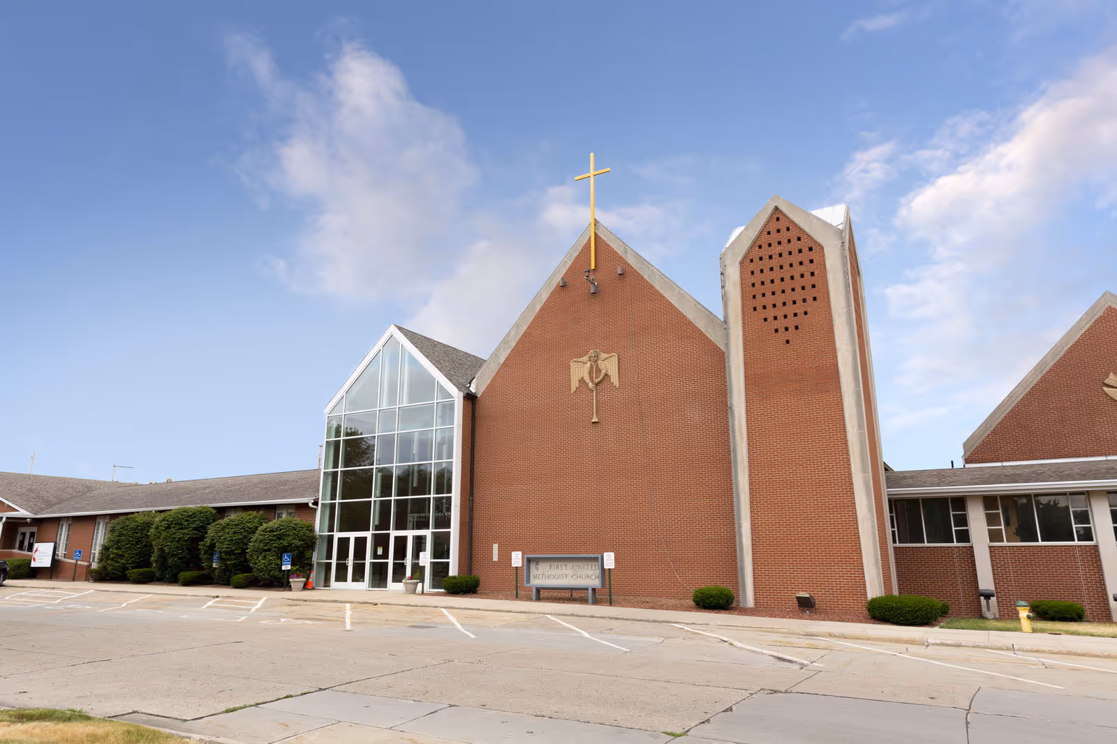 Exterior view of a brick building with a large glass entrance and a tall steeple featuring a cross on top, under a partly cloudy sky.