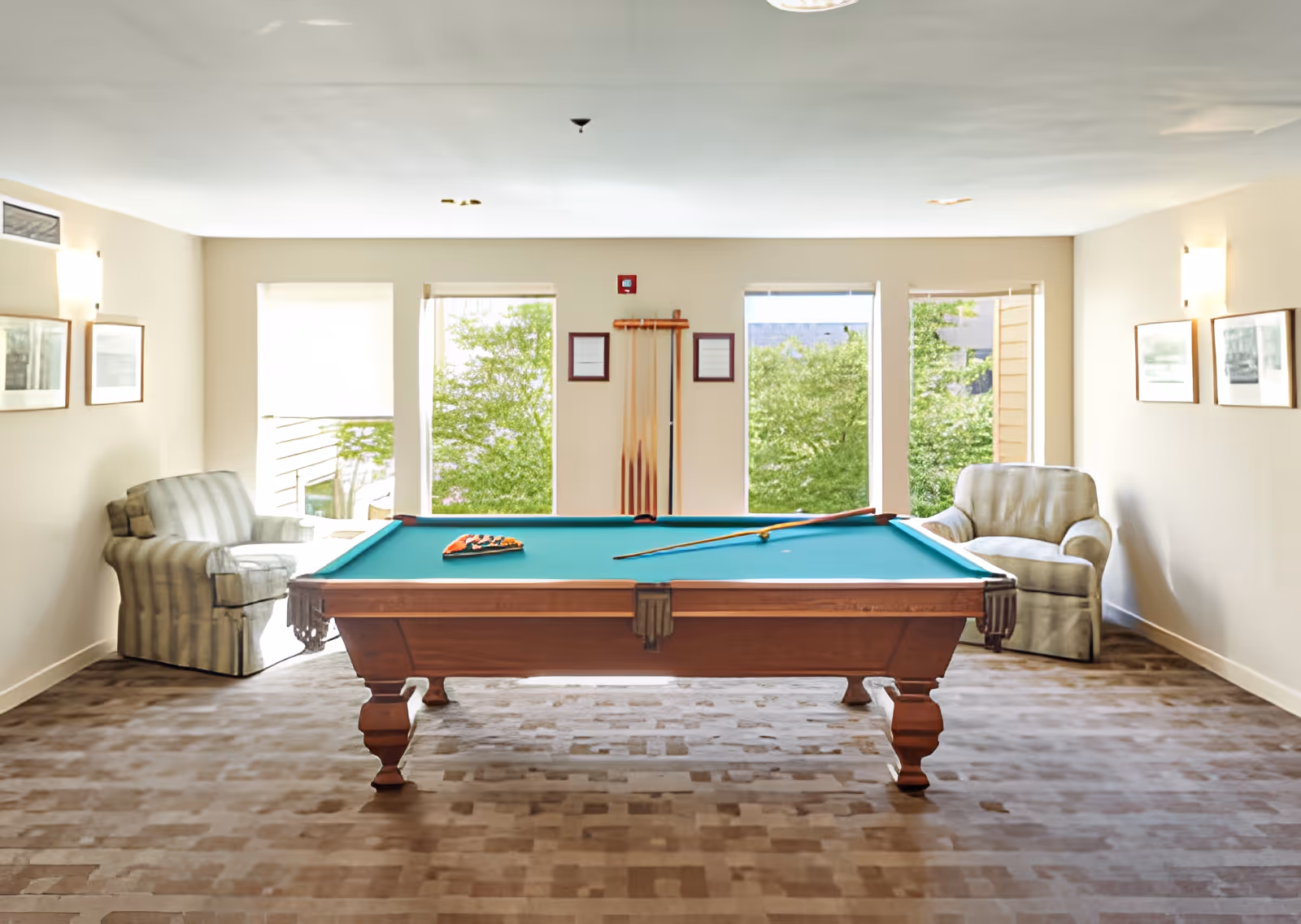 A bright room with a pool table in the center, two armchairs on either side, three large windows showing green trees outside, and pool cues mounted on the wall.
