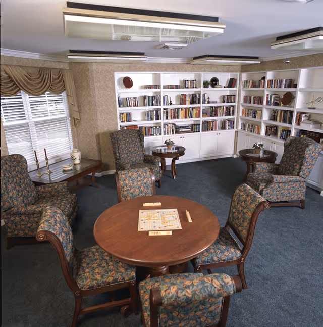 A cozy common room with floral upholstered armchairs and wooden chairs arranged around a round wooden table with a Scrabble board game set up. The room has built-in white bookshelves filled with books and decorative items, a window with beige curtains, and a side table with candles and a vase.
