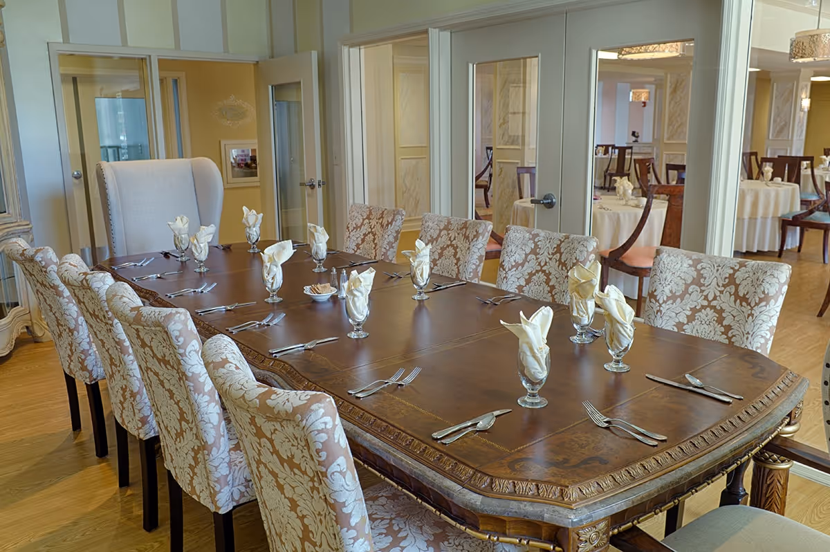 A formal dining room with a large wooden table set for twelve people. The table is adorned with folded white napkins in glass holders and silverware. The chairs around the table have a beige and white floral pattern. In the background, there are glass doors and additional dining tables with similar settings.