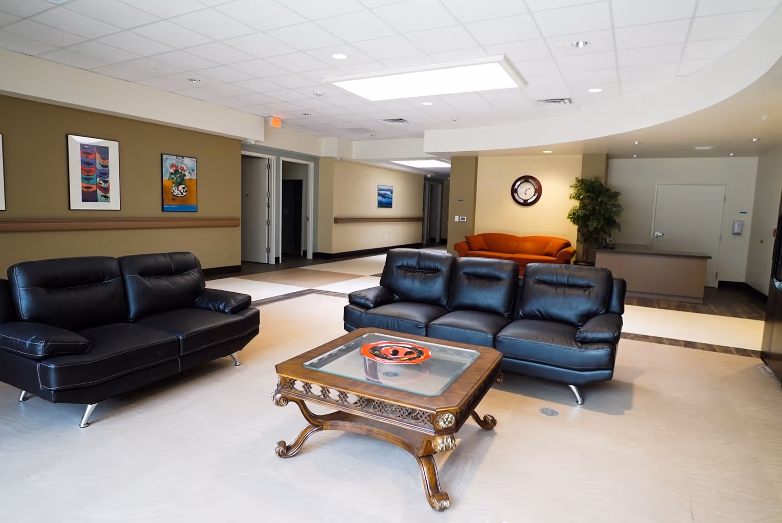 A spacious common area in a senior living facility with two black leather sofas and one orange sofa arranged around a wooden coffee table with a glass top and a decorative bowl. The walls are beige with framed artwork, a clock, and a potted plant. The ceiling has recessed lighting and a large rectangular light panel.