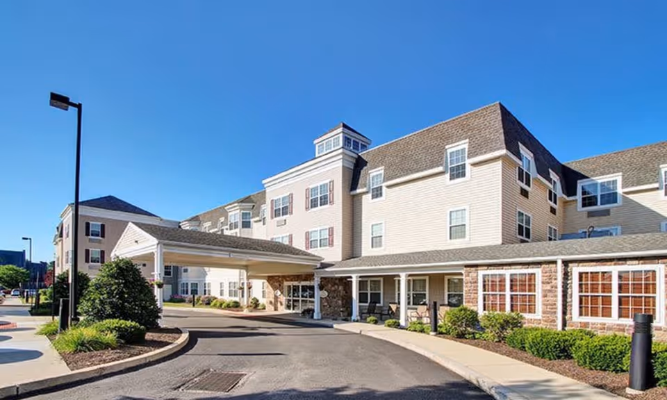 Front exterior of a multi-story senior living building with a covered entry driveway and landscaping under a clear blue sky.