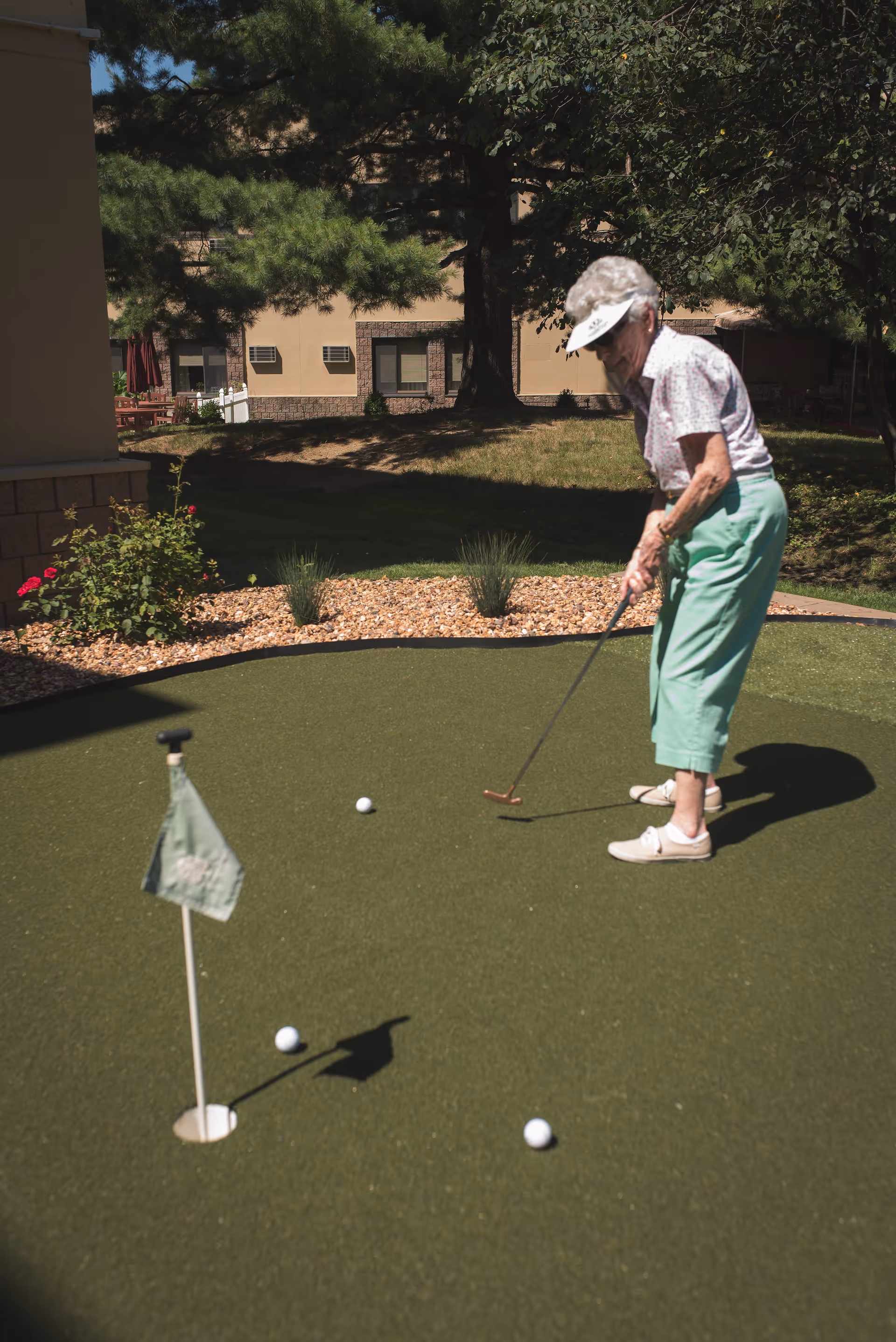 An elderly woman wearing a visor, floral shirt, and green pants is playing mini golf on a putting green outdoors. There are three golf balls on the green near a hole with a small flag. Trees and a building are visible in the background.