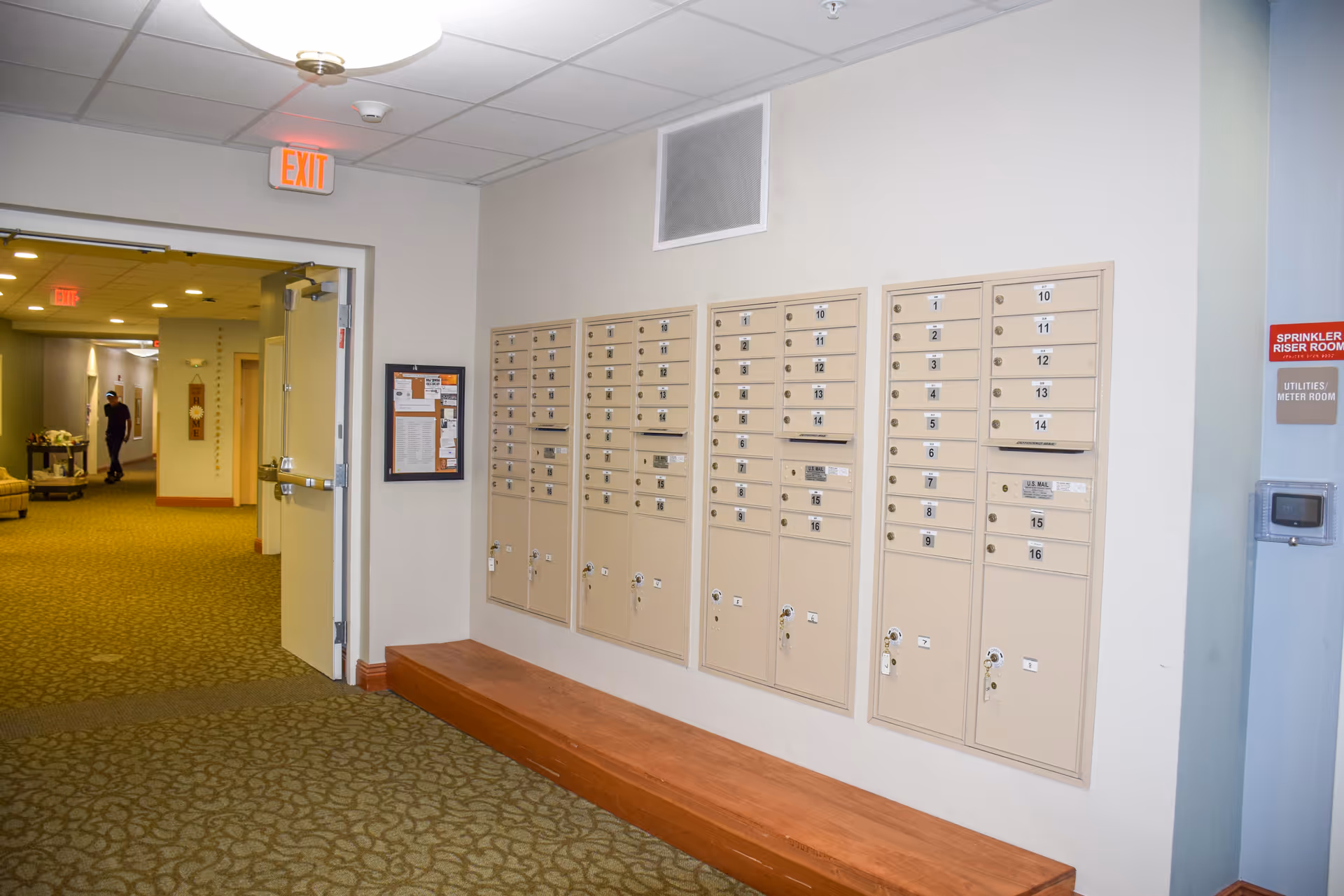 Interior hallway of a senior living facility with a row of beige mailboxes mounted on the wall above a wooden bench. The hallway has carpeted floors and white walls, with an open door leading to a corridor where a person is walking. Exit signs are visible on the ceiling, and there are signs indicating a sprinkler riser room and utilities/meter room on the wall.