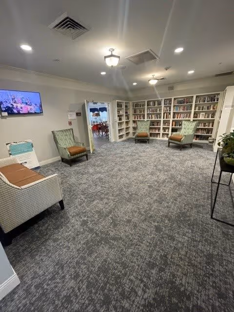 Carpeted communal library lounge with bookshelves along the back wall, several armchairs, a wall-mounted TV, and overhead lights.