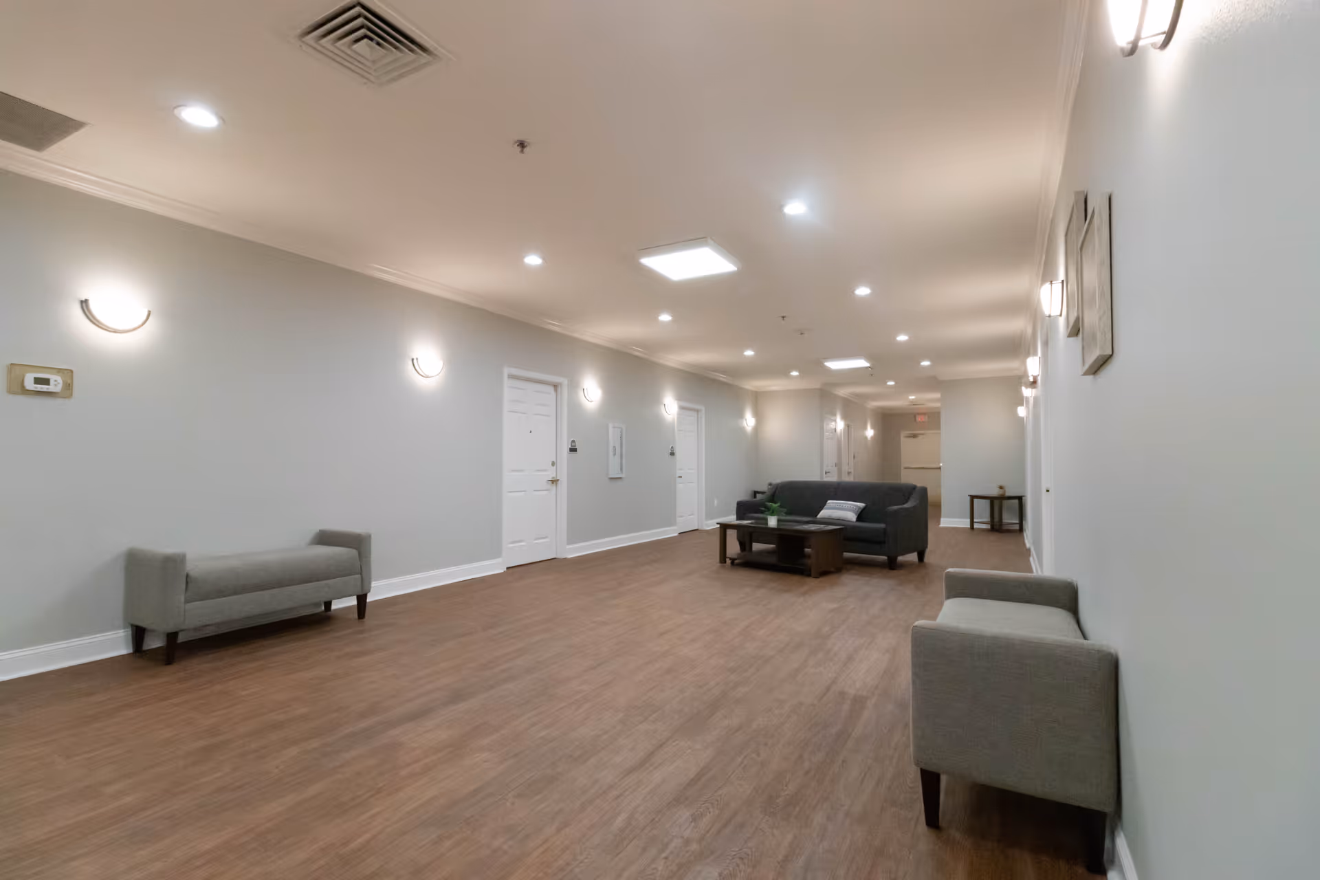 A long hallway with light gray walls and wooden flooring, featuring several white doors along the walls. The hallway is furnished with a gray sofa, a coffee table with a small plant, and two gray upholstered benches. The ceiling has recessed lighting and wall-mounted sconces providing illumination.